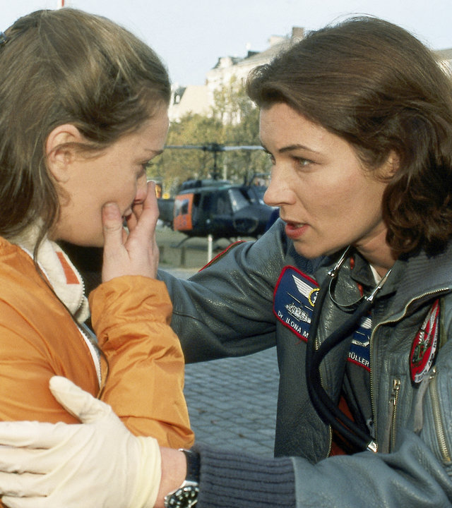 "Die Rettungsflieger - Explosiv": Straßenszene. Im Hintergrund stehen der Rettungshubschrauber und Polizeifahrzeuge. Andrea (Natalie Spinel) und Ilona (Julia Heinemann) stehen sich gegenüber. Ilona neigt sich Andrea zu und spricht beruhigend auf sie ein. Andrea hält sich entsetzt die Hand vor den Mund.