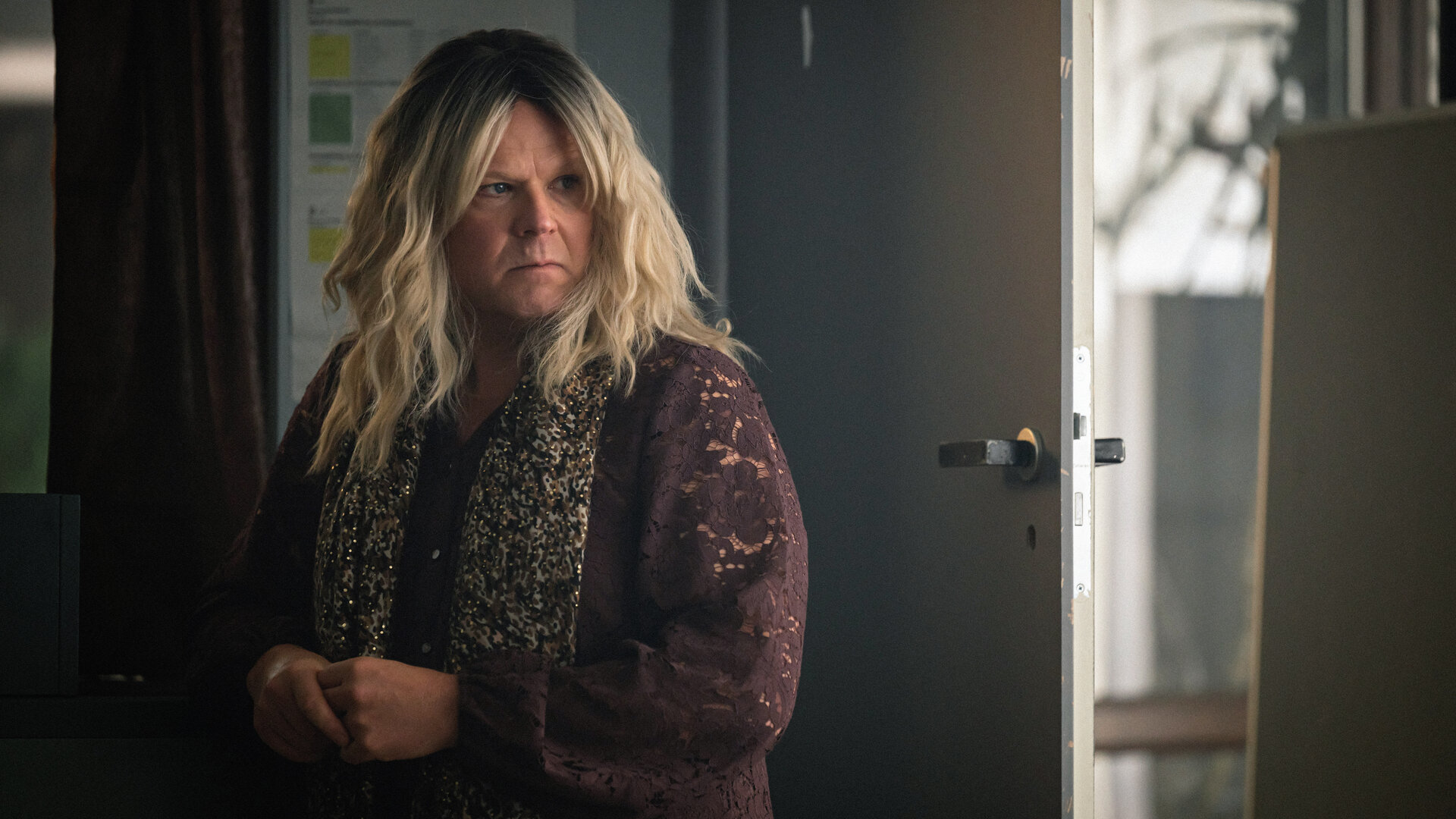 "Toxic Tom - Berit, die heißeste Braut in Oslo": Berit (Anders Baasmo) steht in einer braunen Bluse mit Blumenmuster, mit einem Schal um die Schultern und einer blonden Perücke versonnen in einem Zimmer vor einer geöffneten Türe.