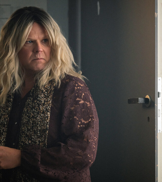 "Toxic Tom - Berit, die heißeste Braut in Oslo": Berit (Anders Baasmo) steht in einer braunen Bluse mit Blumenmuster, mit einem Schal um die Schultern und einer blonden Perücke versonnen in einem Zimmer vor einer geöffneten Türe.