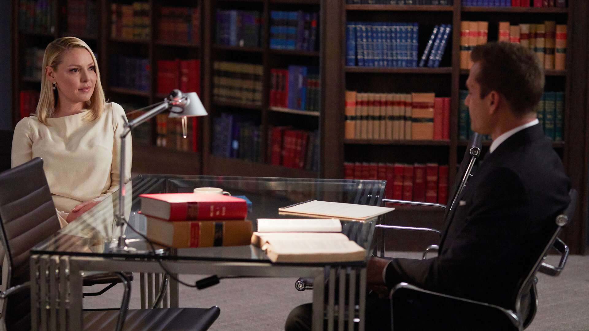 "Suits - Verletzter Stolz": Harvey (Gabriel Macht) und Samantha (Katherine Heigl) sitzen sich an einem Glastisch gegenüber. Sie schauen sich erwartungsvoll an. Im Hintergrund sind gefüllte Bücherregale zu erkennen.