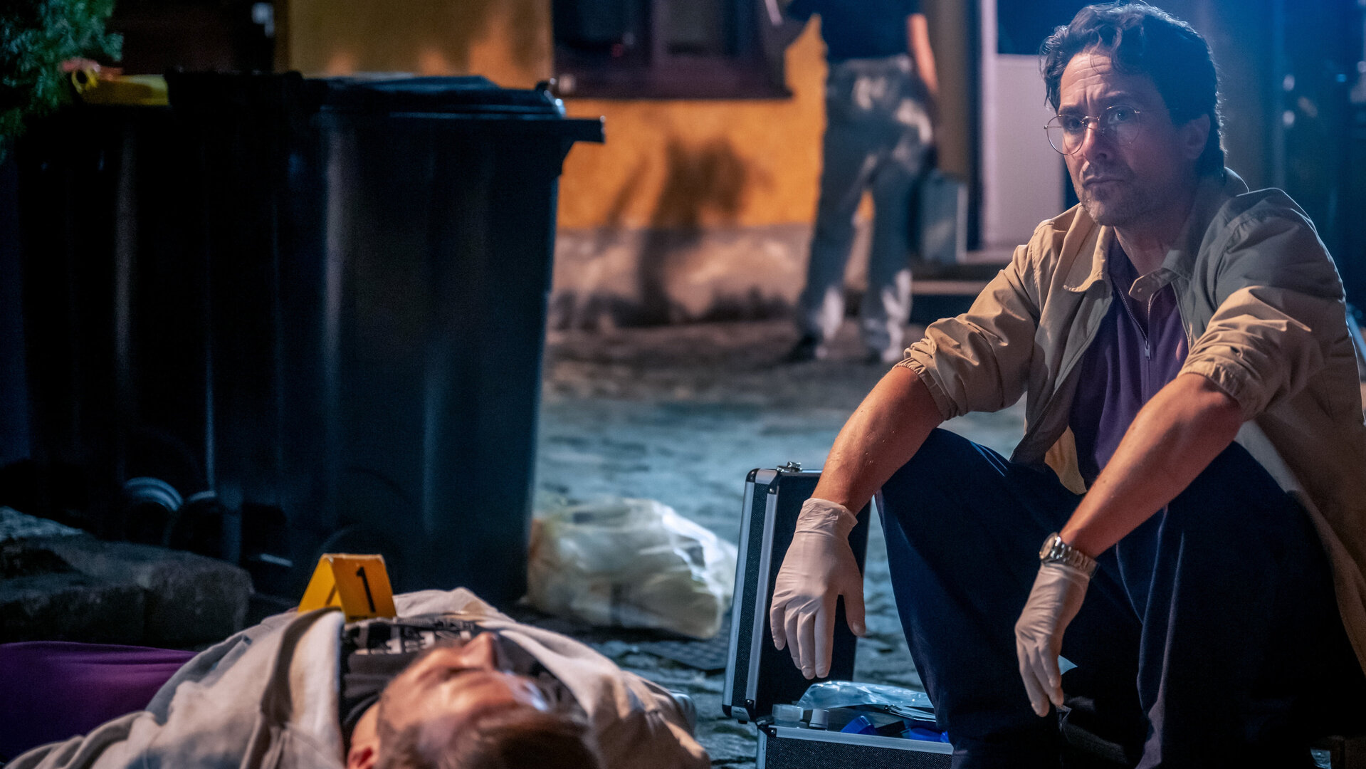 "SOKO Linz - Das Phantom": Gerichtsmediziner Richie (Alexander Pschill) hockt nachdenklich am Tatort. Neben ihm liegt die Leiche von Roman Illbruck (Christoph Bittenauer). Hinter ihnen stehen Mülltonnen und daneben ein Mitarbeiter der Spurensicherung (Komparse) vor einem Haus.