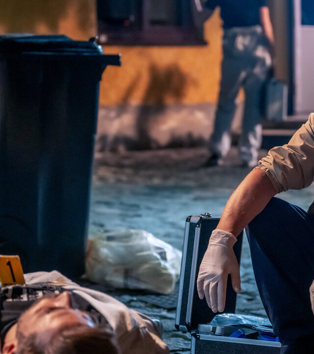 "SOKO Linz - Das Phantom": Gerichtsmediziner Richie (Alexander Pschill) hockt nachdenklich am Tatort. Neben ihm liegt die Leiche von Roman Illbruck (Christoph Bittenauer). Hinter ihnen stehen Mülltonnen und daneben ein Mitarbeiter der Spurensicherung (Komparse) vor einem Haus.