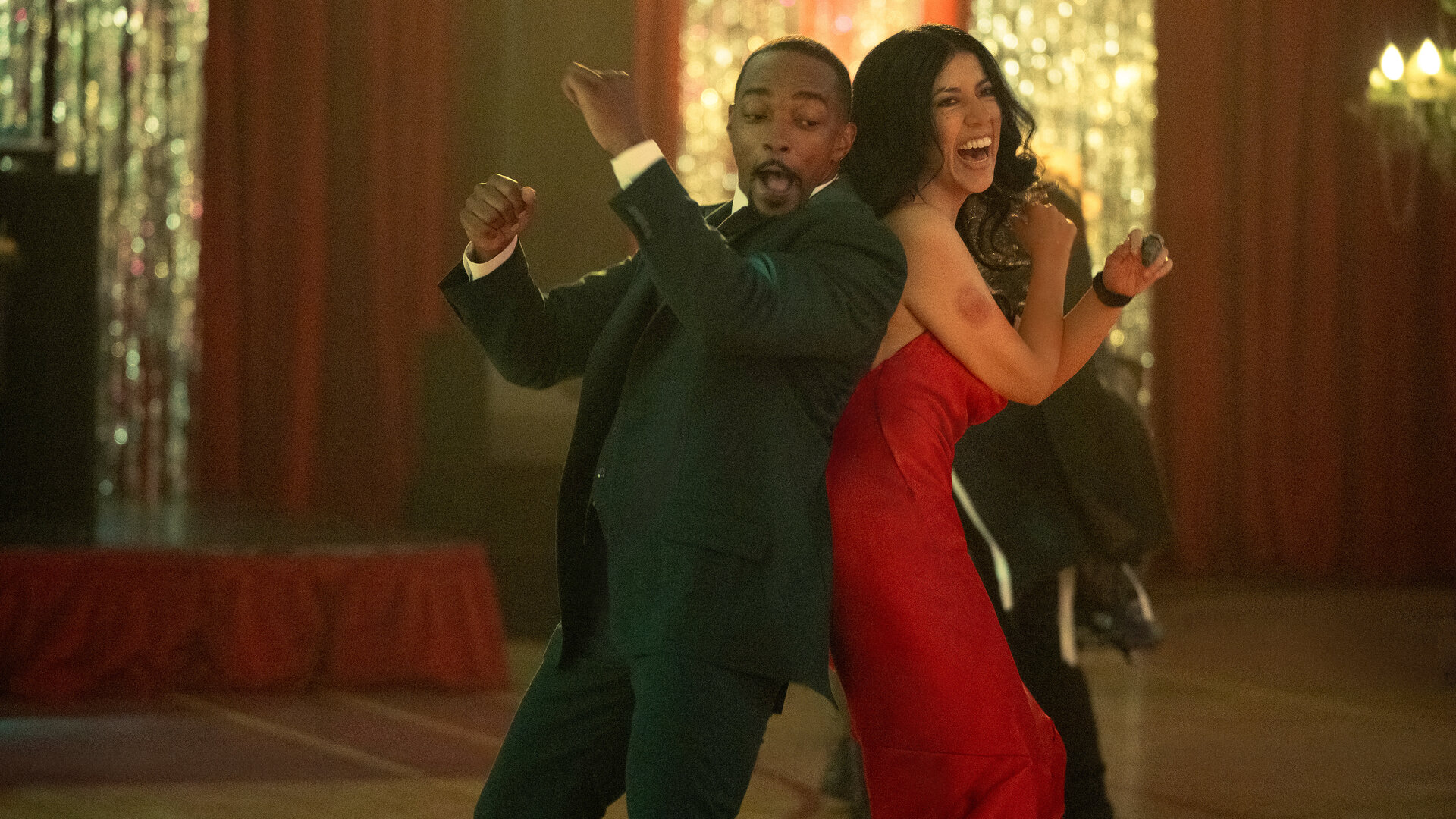 "Twisted Metal - VAVAVUM": In einer festlich geschmückten Sporthalle tanzen John (Anthony Mackie) und Quiet (Stephanie Beatriz) gemeinsam. Sie stehen Rücken an Rücken und werfen die Arme nach oben. John trägt einen schwarzen Anzug mit einem weißen Hemd. Quiet trägt ein rotes Abendkleid mit dünnen Trägern, während ihr dunkles Haar offen über ihre Schultern fällt. John zieht eine Grimasse, während Quiet lacht.