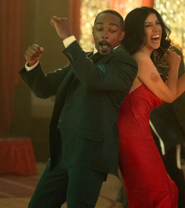 "Twisted Metal - VAVAVUM": In einer festlich geschmückten Sporthalle tanzen John (Anthony Mackie) und Quiet (Stephanie Beatriz) gemeinsam. Sie stehen Rücken an Rücken und werfen die Arme nach oben. John trägt einen schwarzen Anzug mit einem weißen Hemd. Quiet trägt ein rotes Abendkleid mit dünnen Trägern, während ihr dunkles Haar offen über ihre Schultern fällt. John zieht eine Grimasse, während Quiet lacht.