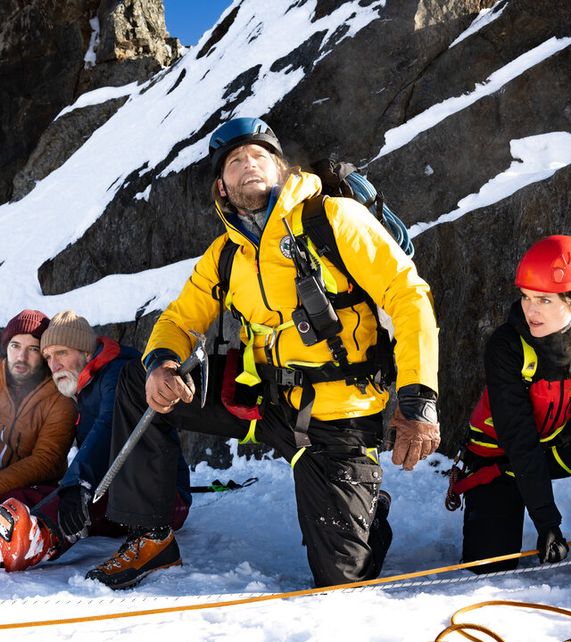 "Die Bergretter: Freier Fall": Im Hochgebirge: Vor einer schneebedeckten Felswand sitzen Hermann Kundig (Jochen Nickel) und Benedikt Kundig (Manuel Mairhofer) nebeneinander im Schnee. Sie wirken geschockt. Im Vordergrund kniet Bergretter Markus Kofler (Sebastian StrÃ¶bel) und blickt Richtung Himmel. Neben ihm kniet seine Kollegin Nina (Josefin Busch). Beide tragen ihre SicherheitsausrÃ¼stung mit Helmen. Vor ihnen sind Rettungsseile ausgelegt.
