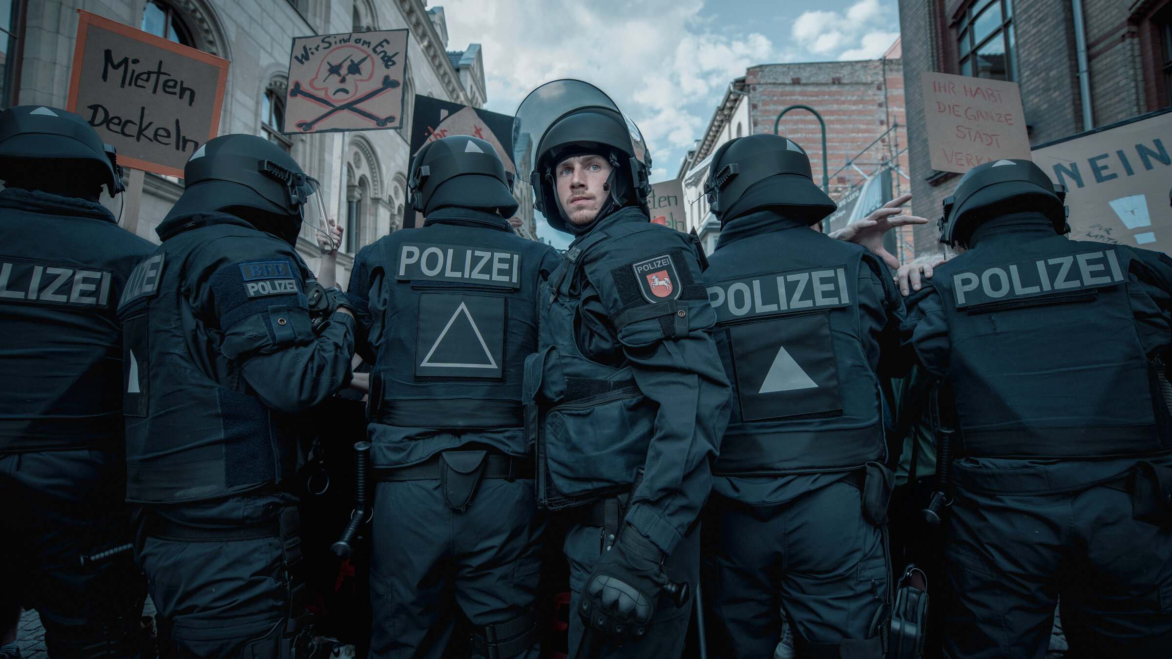"Allein zwischen den Fronten": Jan Vogt (Justus Johanssen) steht vor einem mit schwarzer Einsatzkleidung und Helmen bestückten Einsatztrupp der Polizei und blickt nach vorne in die Kamera. Über den Personen sind Demonstrationsschilder und einige Hausfassaden zu erkennen.