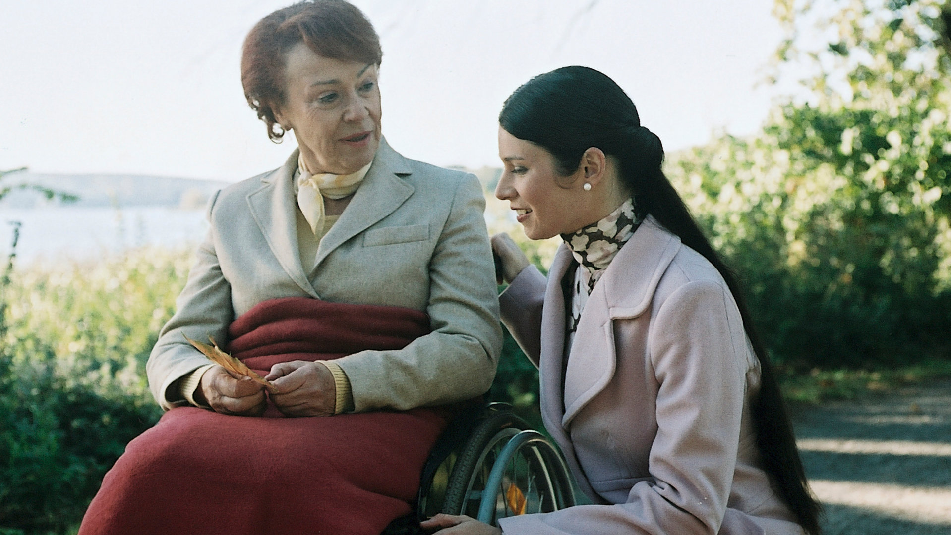 "Bianca - Wege zum Glück: Folge 49": Spaziergang im Park: Judith (Elisabeth Suttrlüty) kniet neben Oma Ursula (Annemone Haas), die im Rollstuhl sitzt und freundlich zu Judith schaut.