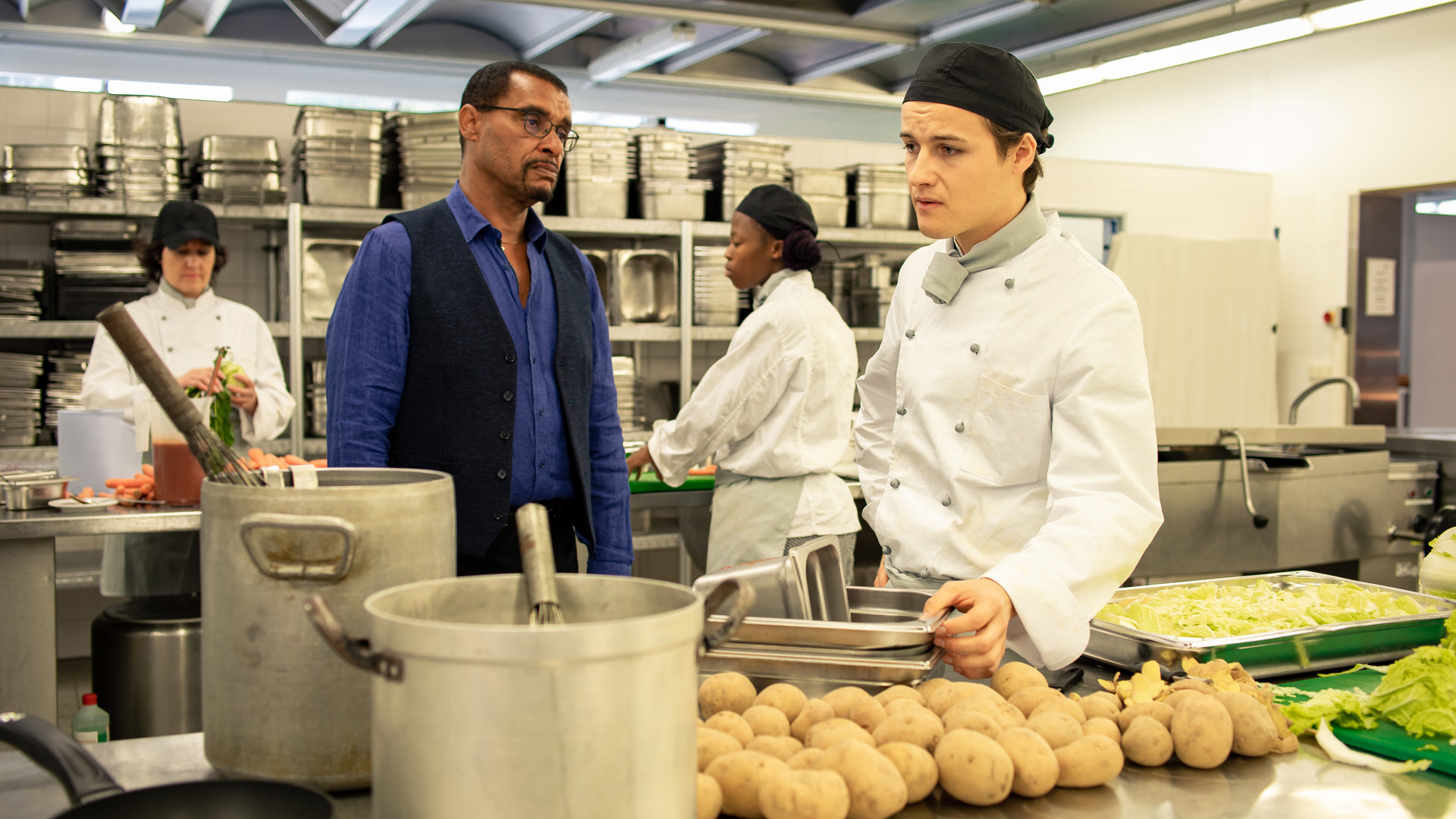 "SOKO München - Tod eines Kochs": Franz Ainfachnur (Christofer v. Beau) befindet sich mit Daniel Steeger (Juri Senft) in einer Großküche. Der Kommissar sieht den jungen Koch ernst an. Daniel Steeger steht neben ihm an einem Stahltisch und blickt bedrückt zur Seite. Er trägt ein weiße Kochjacke und einen Kopfschutz. Der Koch greift nach Edelstahlbehältern, die vor ihm auf dem Tisch stehen. Außerdem liegen dort Kartoffeln, zwei große Kochtöpfe stehen an der Seite und und neben ihm steht ein Behälter mit Salat. Im Hintergrund sieht man zwei weitere Köchinnen bei der Arbeit.