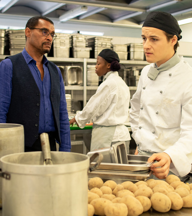 "SOKO München - Tod eines Kochs": Franz Ainfachnur (Christofer v. Beau) befindet sich mit Daniel Steeger (Juri Senft) in einer Großküche. Der Kommissar sieht den jungen Koch ernst an. Daniel Steeger steht neben ihm an einem Stahltisch und blickt bedrückt zur Seite. Er trägt ein weiße Kochjacke und einen Kopfschutz. Der Koch greift nach Edelstahlbehältern, die vor ihm auf dem Tisch stehen. Außerdem liegen dort Kartoffeln, zwei große Kochtöpfe stehen an der Seite und und neben ihm steht ein Behälter mit Salat. Im Hintergrund sieht man zwei weitere Köchinnen bei der Arbeit.