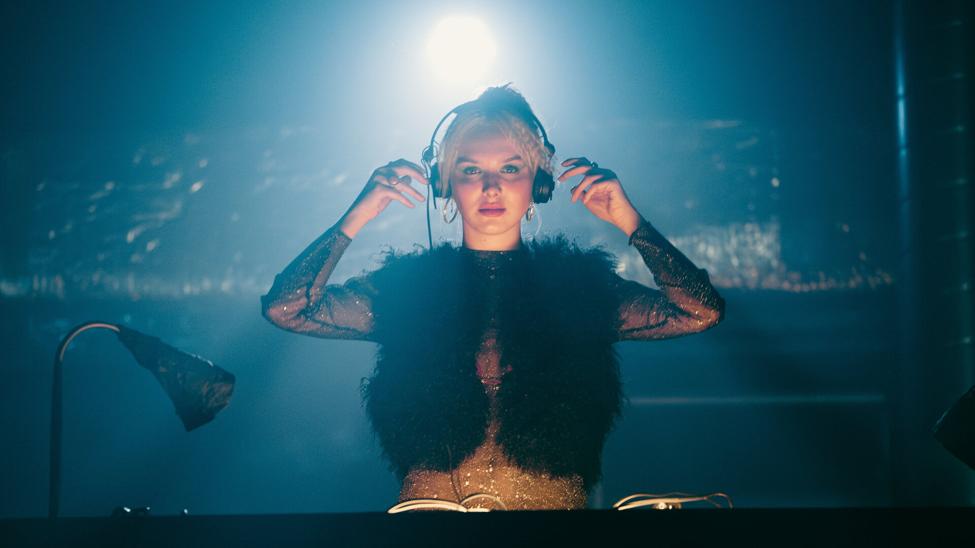 "München Beats - Folge 4": Linda Bierwirth (Jule Hermann) steht hinter einem DJ-Pult und blickt in die Kamera. Sie steht vor dunklem Hintergrund, trägt Kopfhörer und wird von einer Leuchte, die über ihr hängt, angestrahlt.