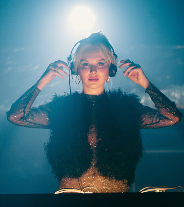 "München Beats - Folge 4": Linda Bierwirth (Jule Hermann) steht hinter einem DJ-Pult und blickt in die Kamera. Sie steht vor dunklem Hintergrund, trägt Kopfhörer und wird von einer Leuchte, die über ihr hängt, angestrahlt.