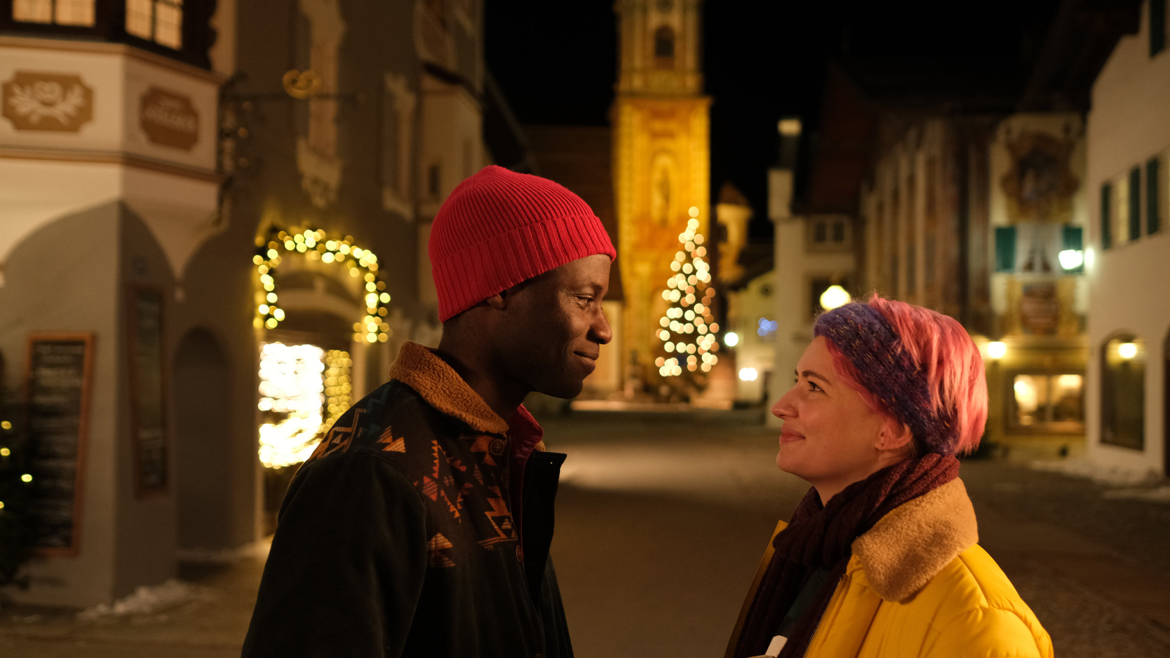 „Weihnachtspäckchen… haben alle zu tragen“: Adika (Yann Mbiene) und Tessa (Zoë Valks) stehen sich auf einer nächtlichen Straße gegenüber und lächeln sich verliebt an. Im Hintergrund steht eine beleuchtete Kirche, ein großer geschmückter Weihnachtsbaum sowie Häuser mit weihnachtlich beleuchteten Schaufenstern.