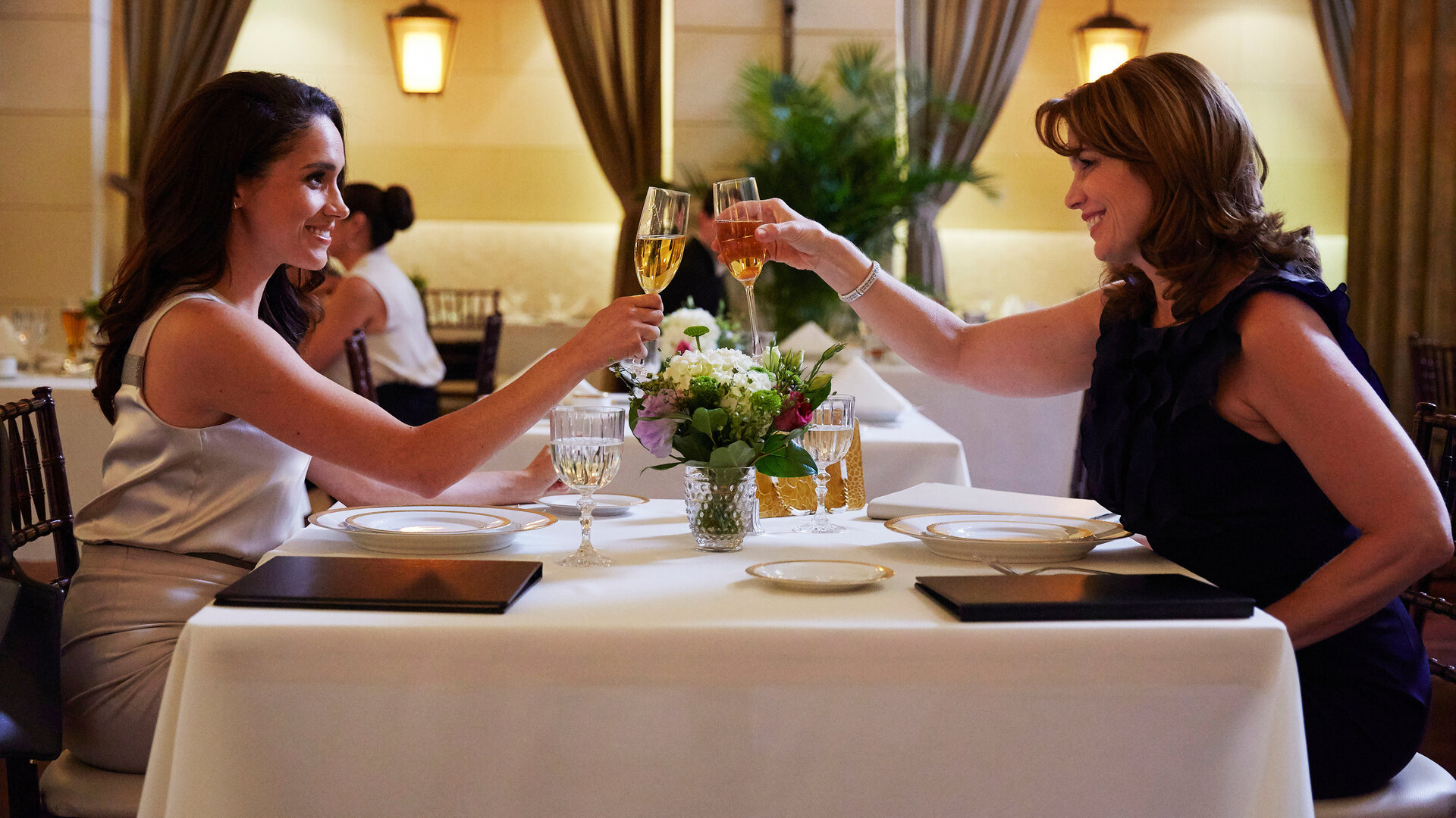 "Suits - Ungebetene Gäste": Rachel (Meghan Markle) und ihre Mutter Laura (Megan Gallagher) sitzen an einem festlich gedeckten Tisch in einem noblen Restaurant und stoßen einander zulächelnd mit Champagnergläsern an.