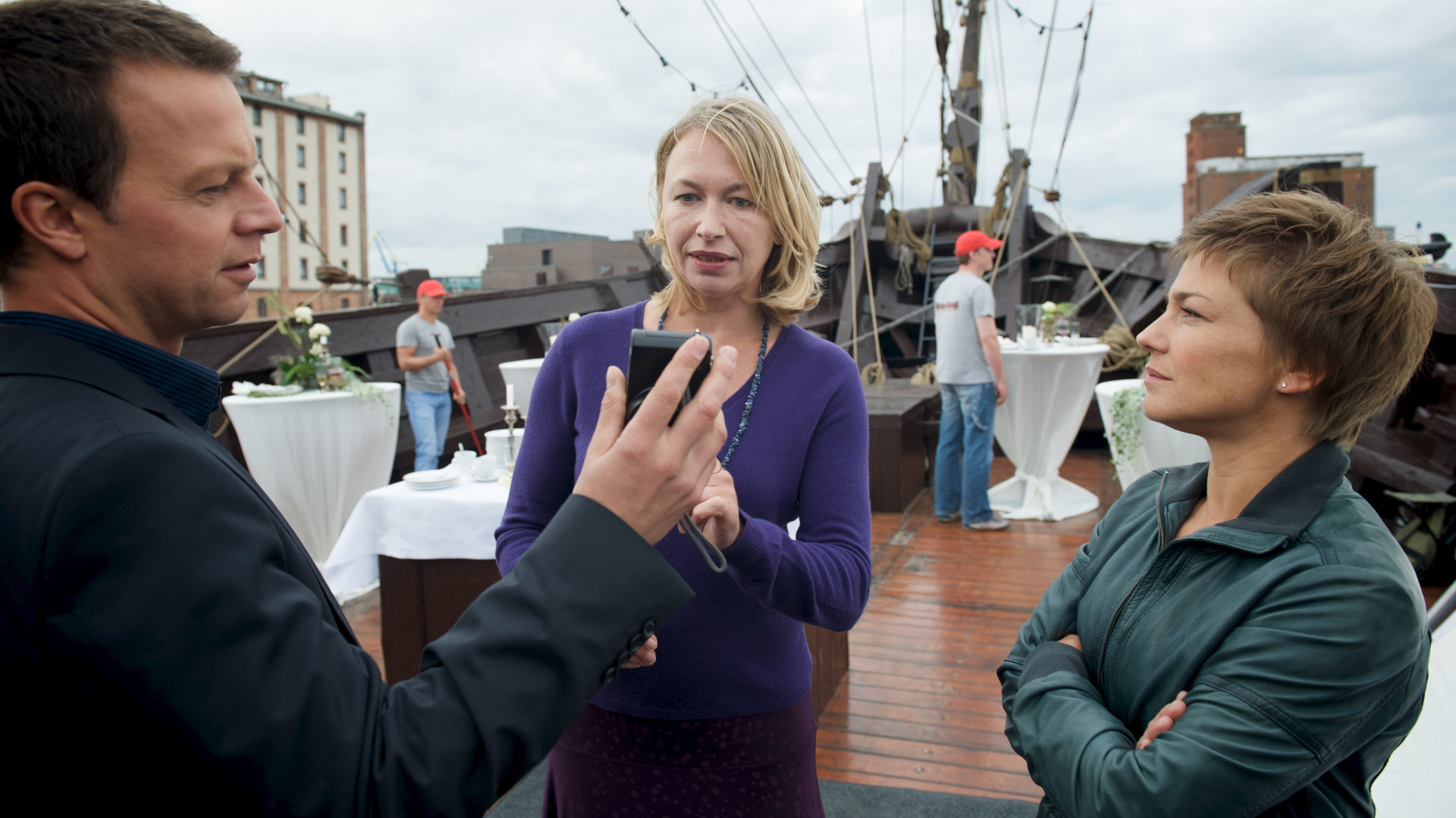 "SOKO Wismar - Ein Stich ins Herz": Auf einem Schiffsdeck, auf dem noch Stehtische für eine Feierlichkeit stehen, zeigt Sven Herzog (Michael Härle) Sandra Bosse (Therese Hämer) ein Bild auf dem Smartphone. Katrin Börensen (Claudia Schmutzler) steht dabei.