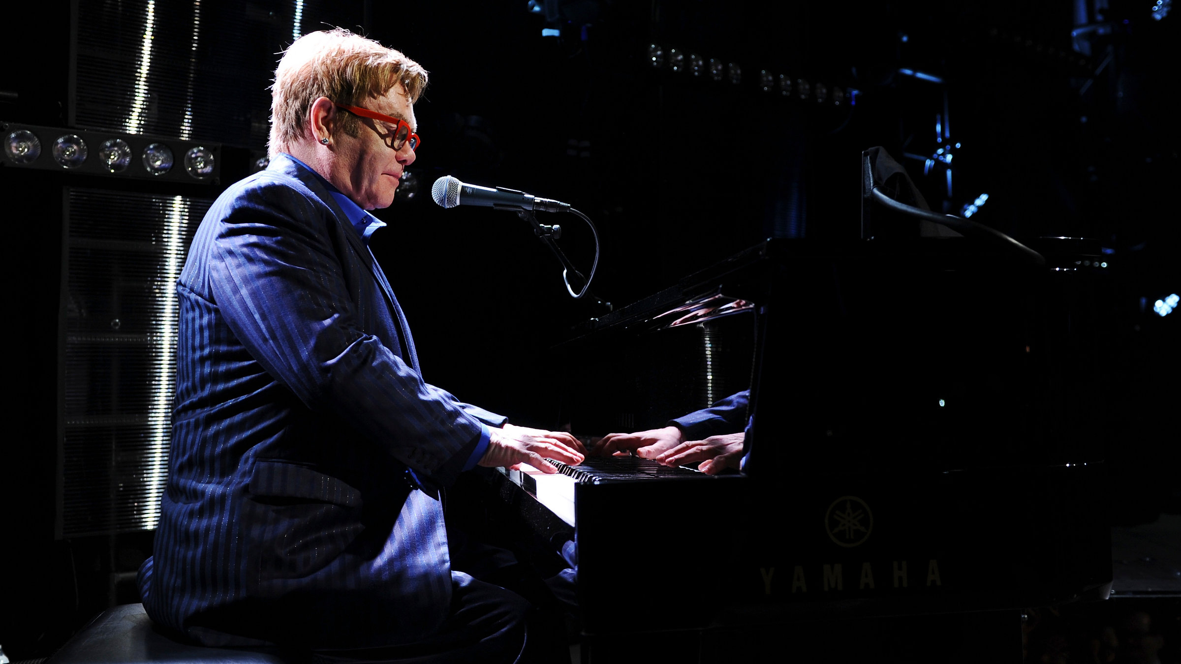 "The True Story of Elton John": Elton John trägt eine rote Sonnenbrille und ein gestreiftes blaues Sakko. Er spielt Klavier.