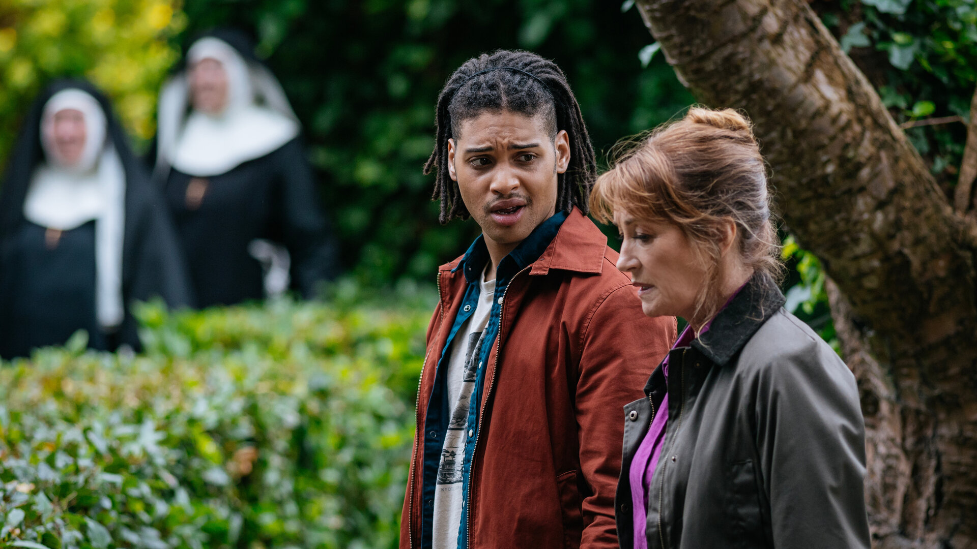 "Harry Wild – Mörderjagd in Dublin - Das Kloster der schweigenden Nonnen": Fergus Reid (Rohan Nedd) und Harry Wild (Jane Seymour) stehen nebeneinander vor einem Baum und einer Hecke. Im Hintergrund sind in der Unschärfe zwei Nonnen zu sehen.