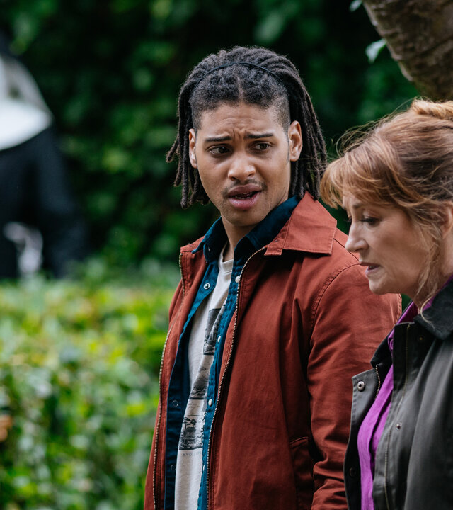 "Harry Wild – Mörderjagd in Dublin - Das Kloster der schweigenden Nonnen": Fergus Reid (Rohan Nedd) und Harry Wild (Jane Seymour) stehen nebeneinander vor einem Baum und einer Hecke. Im Hintergrund sind in der Unschärfe zwei Nonnen zu sehen.