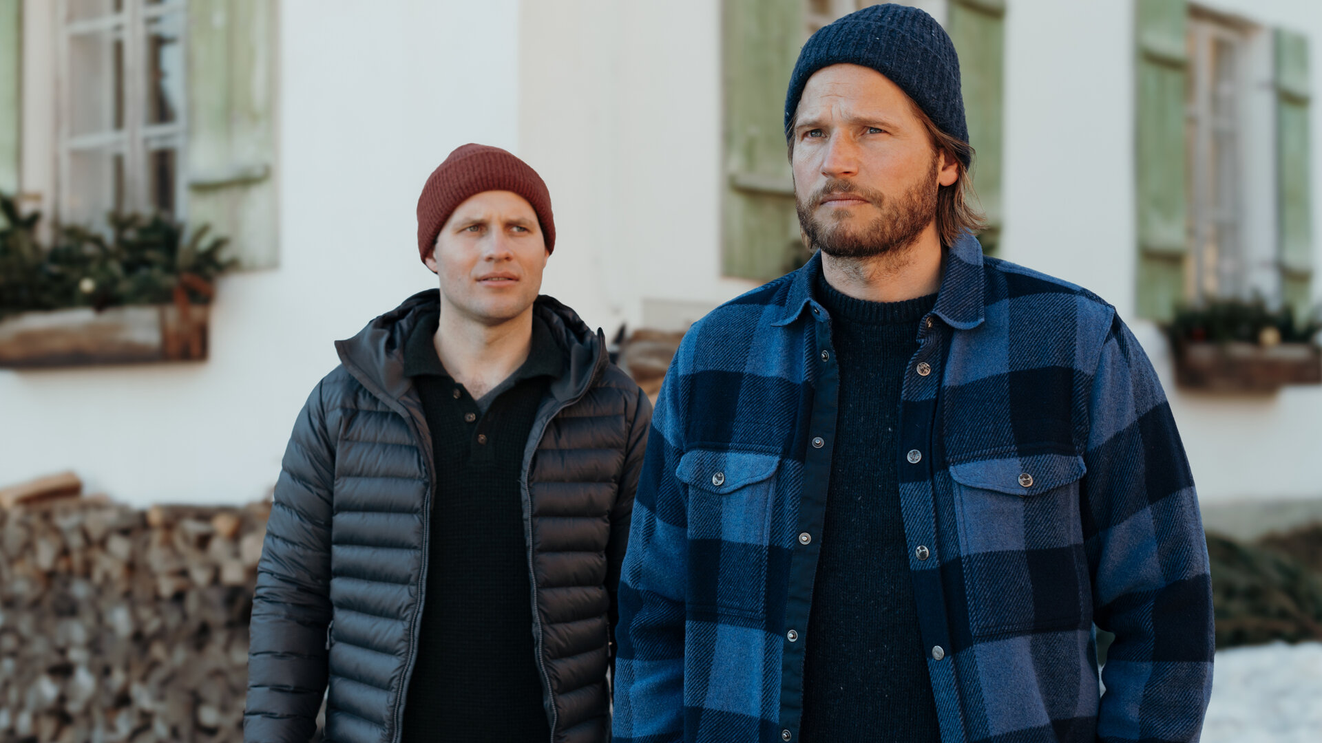 "Die Bergretter: Im Stich gelassen": Auf Emilies Hof: Nachdenklich, die Hände in den Hosentaschen, steht Markus Kofler (Sebastian Ströbel) vor dem Haus. Hinter ihm steht Jan Fugain (Arne Löber) und schaut ihn fragend an. Beide tragen Winterbekleidung und Mützen. Im Hintergrund liegt Schnee und in den Blumentöpfen auf den Fenstersimsen stecken mit Weihnachtskugeln geschmückte Tannenzweige.