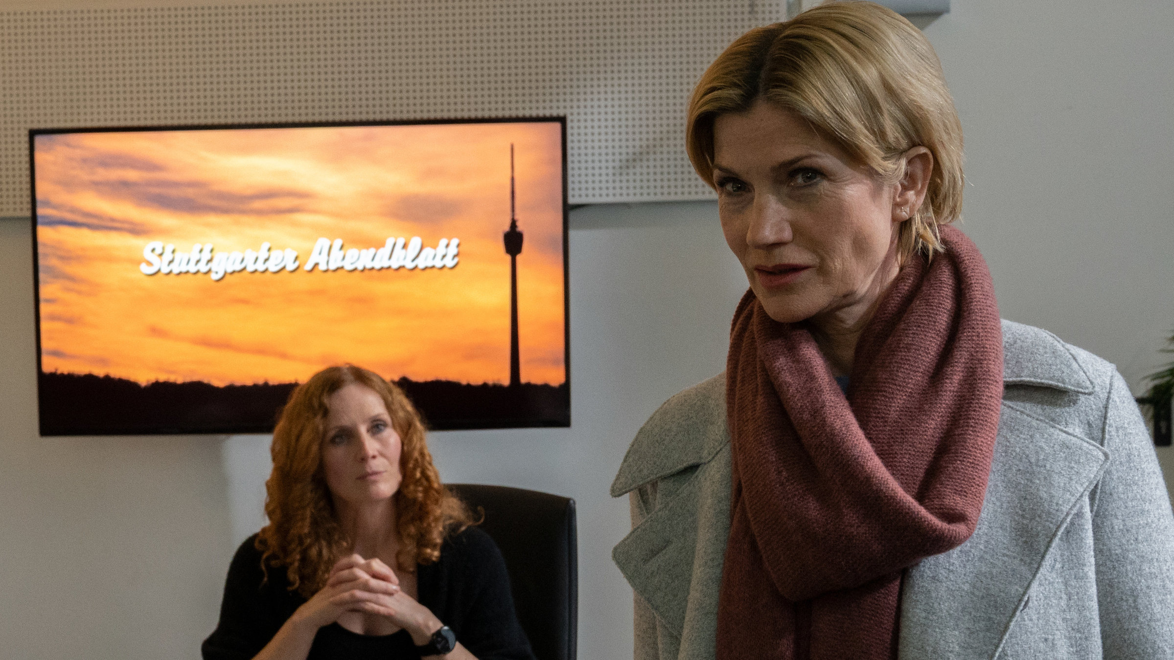 "SOKO Stuttgart – #MeToo": Martina Seiffert (Astrid M. Fünderich) befragt Verlagschefin Elena Krüger (Jutta Fastian) in deren Büro. Krüger sitzt an ihrem Schreibtisch, auf dem zahlreiche Büroutensilien sowie Unterlagen zu sehen sind. An der Wand hinter ihr ist ein Bildschirm angebracht, auf dem der Schriftzug "Stuttgarter Abendblatt" zu sehen ist. Mit gefalteten Händen schaut Krüger mit souveränem Blick auf Martina, die ihr den Rücken zuwendet.