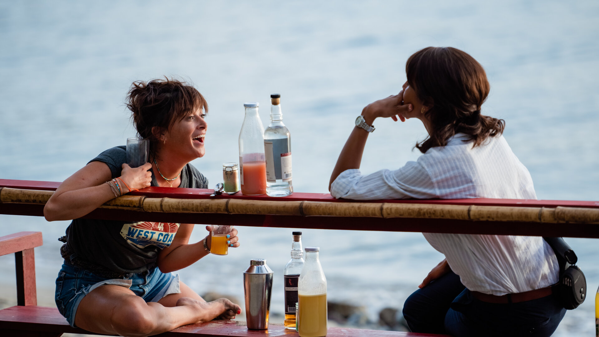 "Deadly Tropics - Undercover": Mélissa (Sonia Rolland) und Gaëlle (Béatrice de la Boulaye) sitzen gemeinsam auf einer Bank am Strand und blicken einander an. Gaëlle lacht dabei und hält in jeder Hand ein Glas. Zwischen ihnen stehen Alkoholflaschen.