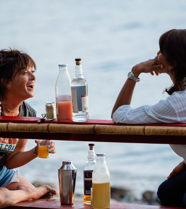 "Deadly Tropics - Undercover": Mélissa (Sonia Rolland) und Gaëlle (Béatrice de la Boulaye) sitzen gemeinsam auf einer Bank am Strand und blicken einander an. Gaëlle lacht dabei und hält in jeder Hand ein Glas. Zwischen ihnen stehen Alkoholflaschen.