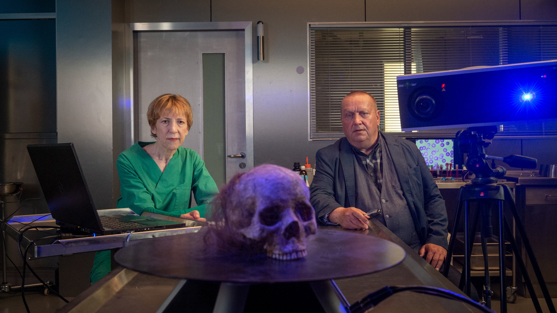 "SOKO Wismar – Der schöne Klaus": Auf einem sich drehenden, runden Podest steht ein weiblicher Totenschädel. Der Schädel ist mit einem lilafarbenen Licht angeleuchtet. Dahinter sitzen Helene und Reuter und blicken konzentriert auf den Schädel. Helene trägt ihren grünen Kittel und hat die Hände zusammen gefaltet. Reuter hat seine Brille abgenommen und trägt sie in der rechten Hand. Im Hintergrund stehen Proben herum, auf einem Bildschirm sind Zellstrukturen stark vergrößert abgebildet.