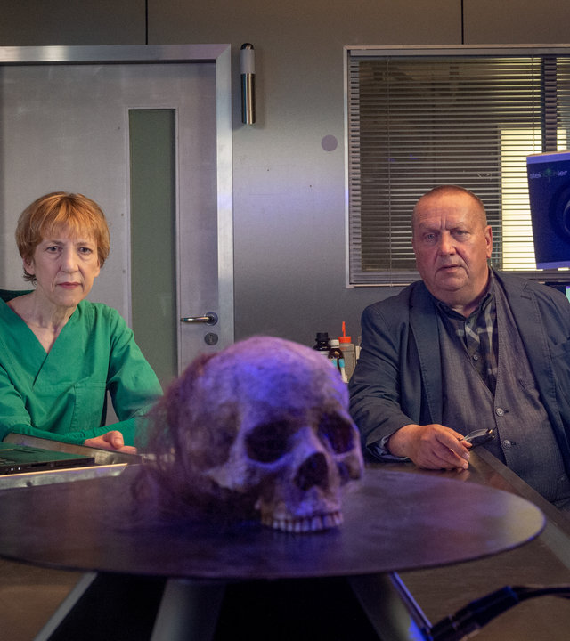 "SOKO Wismar – Der schöne Klaus": Auf einem sich drehenden, runden Podest steht ein weiblicher Totenschädel. Der Schädel ist mit einem lilafarbenen Licht angeleuchtet. Dahinter sitzen Helene und Reuter und blicken konzentriert auf den Schädel. Helene trägt ihren grünen Kittel und hat die Hände zusammen gefaltet. Reuter hat seine Brille abgenommen und trägt sie in der rechten Hand. Im Hintergrund stehen Proben herum, auf einem Bildschirm sind Zellstrukturen stark vergrößert abgebildet.