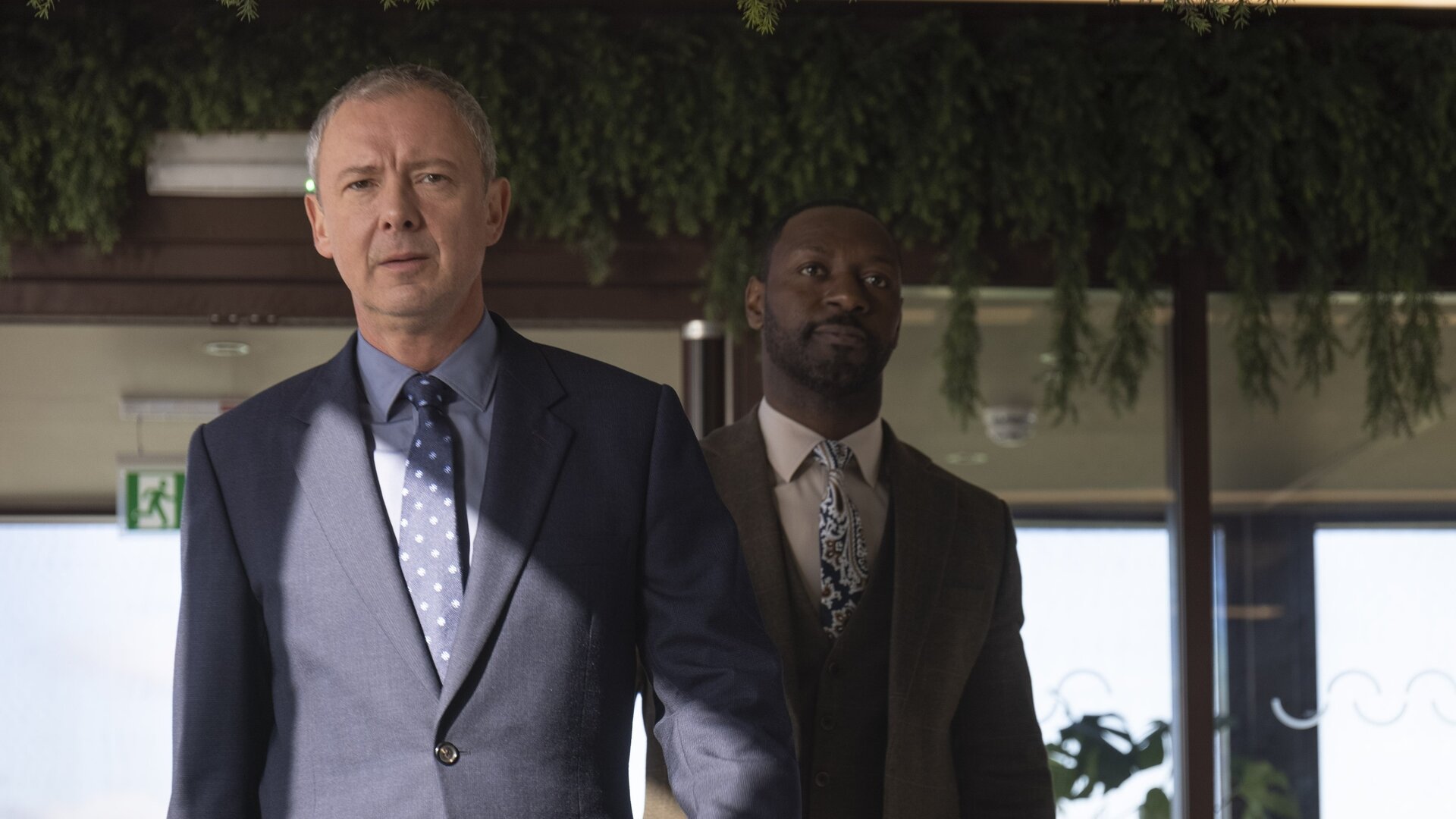 "Detective Grace - So gut wie tot": DS Roy Grace (John Simm) und DS Glenn Branson (Ritchie Campbell) gehen hintereinander durch ein leeres, gehobenes Restaurant. Sie wirken entschlossen.