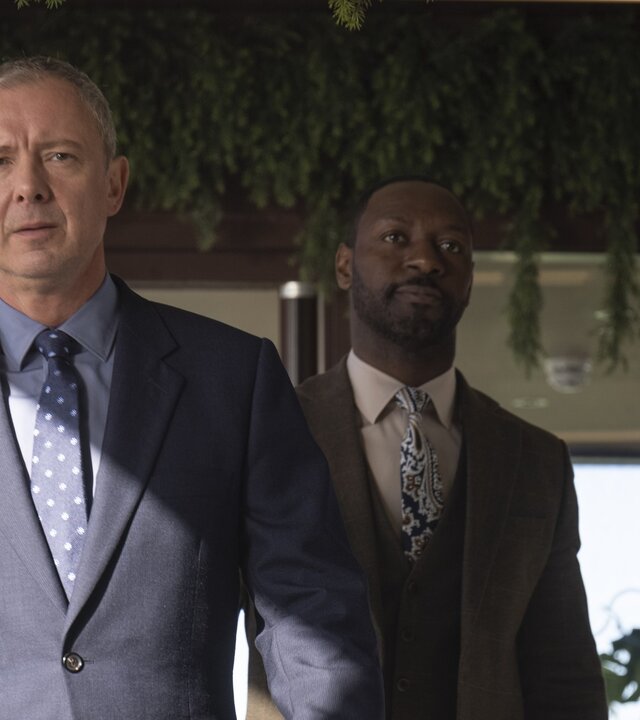 "Detective Grace - So gut wie tot": DS Roy Grace (John Simm) und DS Glenn Branson (Ritchie Campbell) gehen hintereinander durch ein leeres, gehobenes Restaurant. Sie wirken entschlossen.