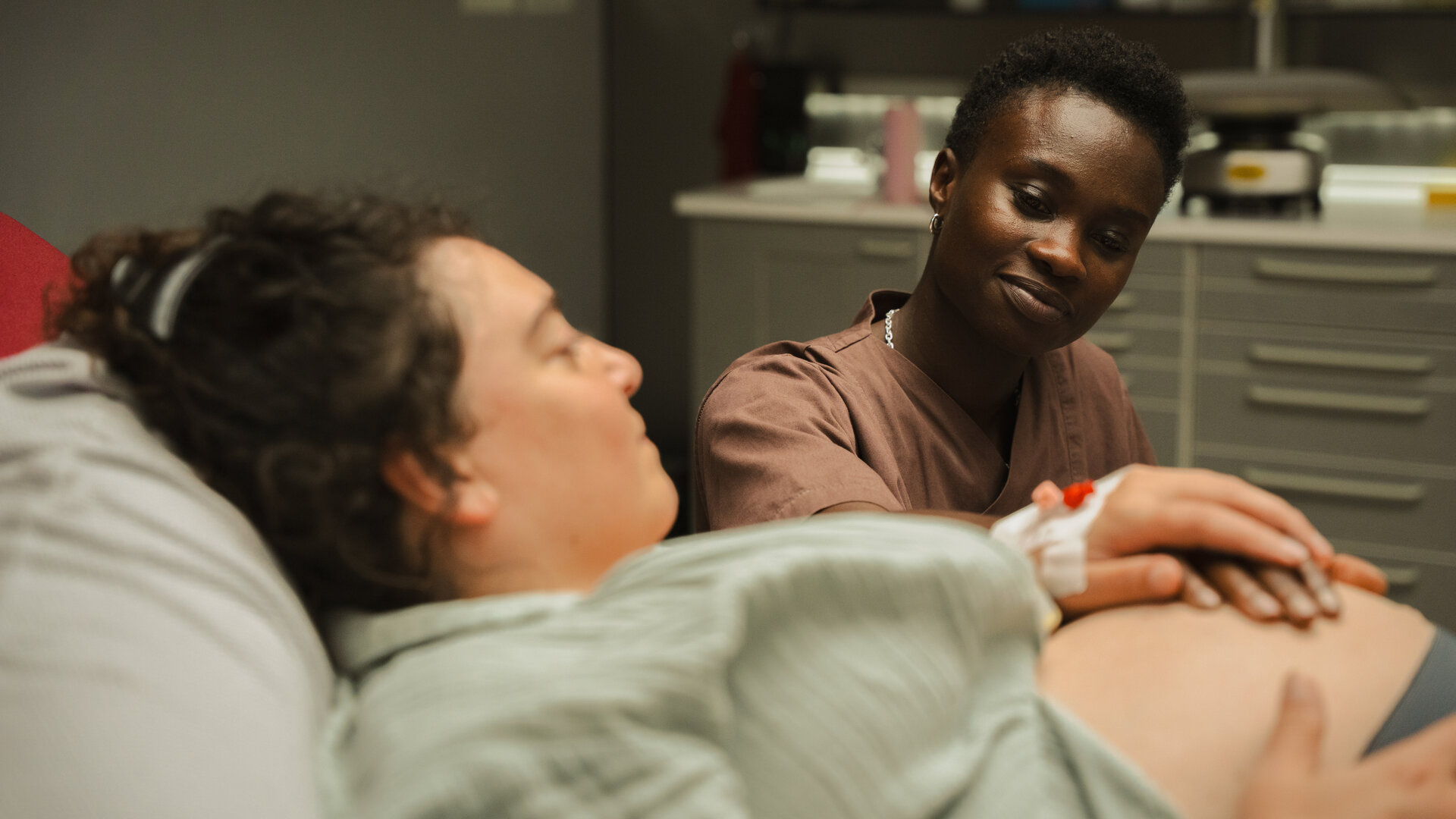"PUSH - Akzeptanz": Sibel (Nuriye Jendroßek) liegt im Patientenbett und schaut zu Tara (Lorna Ishema), die an ihrem Bett sitzt und eine Hand auf Sibels Babybauch legt. Sibel hält Taras Hand.