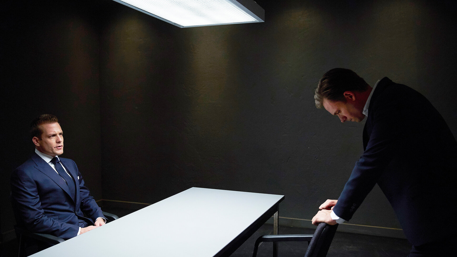 "Suits - Ärger im Anzug": Harvey (Gabriel Macht) sitzt in einem Vernehmungsraum an einem Tisch und schaut Mike (Patrick J. Adams) an, der ihm gegenüber nach vorne gebeugt steht und seine Hände auf einer Armlehne abgelegt hat, wobei er verzweifelt wirkt.