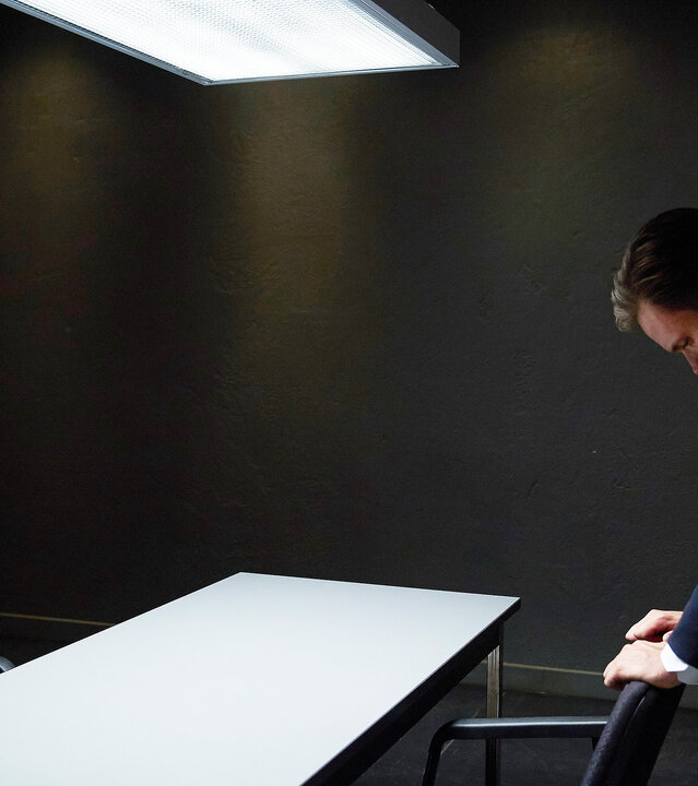 "Suits - Ärger im Anzug": Harvey (Gabriel Macht) sitzt in einem Vernehmungsraum an einem Tisch und schaut Mike (Patrick J. Adams) an, der ihm gegenüber nach vorne gebeugt steht und seine Hände auf einer Armlehne abgelegt hat, wobei er verzweifelt wirkt.