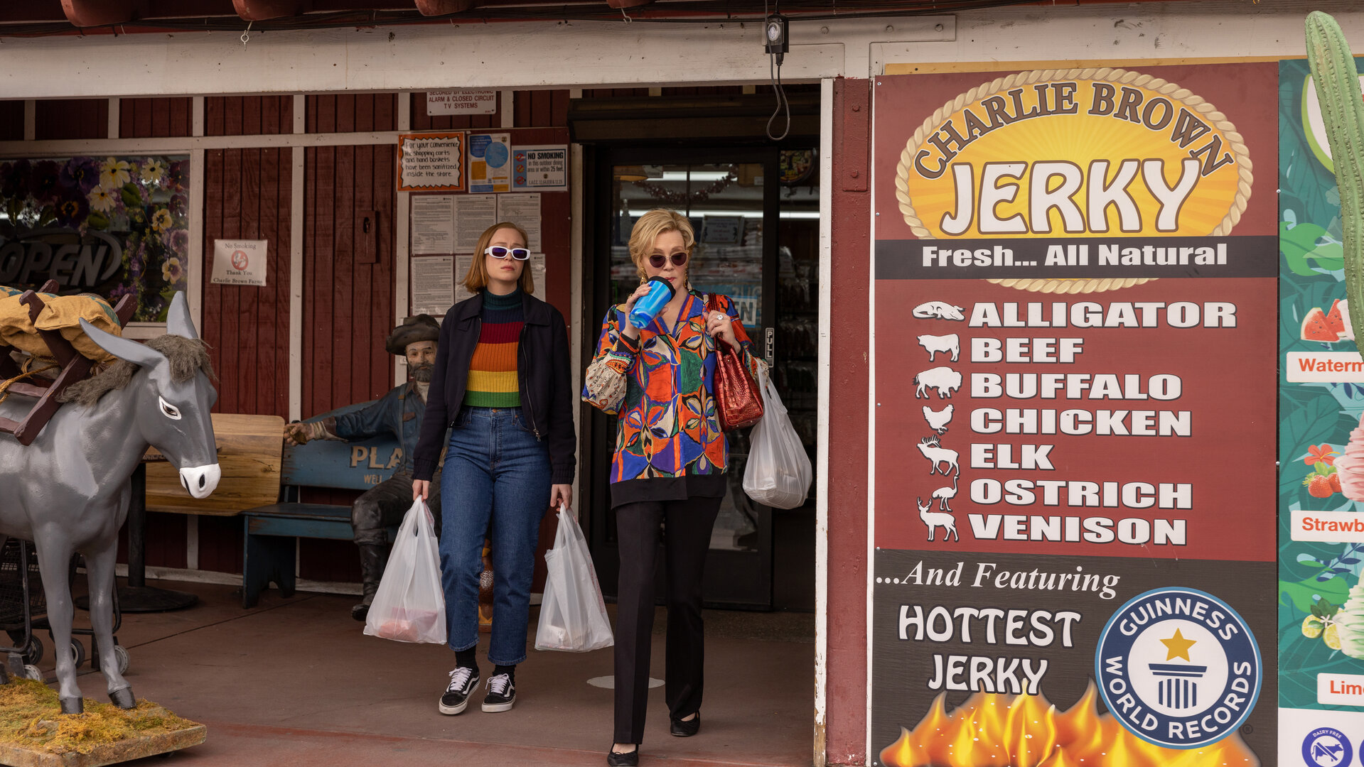 "Hacks - Quid pro quo": (Hannah Einbinder) und Deborah (Jean Smart) tragen Sonnenbrillen und treten mit Einkaufstaschen aus einem Geschäft, während Deborah aus einem Trinkbecher mit Strohhalm trinkt. Neben dem Eingang stehen eine Statue eines beladenen Esels und eine Werbetafel mit Angeboten für verschiedene Fleischsorten.
