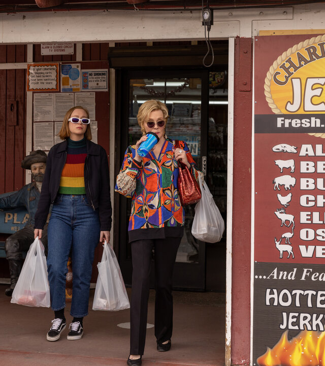 "Hacks - Quid pro quo": (Hannah Einbinder) und Deborah (Jean Smart) tragen Sonnenbrillen und treten mit Einkaufstaschen aus einem Geschäft, während Deborah aus einem Trinkbecher mit Strohhalm trinkt. Neben dem Eingang stehen eine Statue eines beladenen Esels und eine Werbetafel mit Angeboten für verschiedene Fleischsorten.