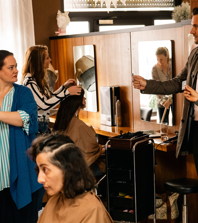 "SOKO Köln - Nur der Moment": Vanessa Haas (Kerstin Landsmann) und Jonas Fischer (Lukas Piloty) stehen in einem Friseursalon nebeneinander und zeigen ihre Ausweise Ada Hoffmann (Katrin Filzen) vor. Diese dreht sich mit ernstem Blick zu ihnen, während sie gerade einer Kundin (Komparsin) Lockenwickler eindreht. Davor sitzt eine weitere Kundin (Komparsin). Hinter Ada befinden sich zwei weitere Frisörinnen (Komparsinnen) bei der Arbeit und eine weitere Kundin (Komparsin) sitzt auf einem Stuhl vor einem Spiegel.