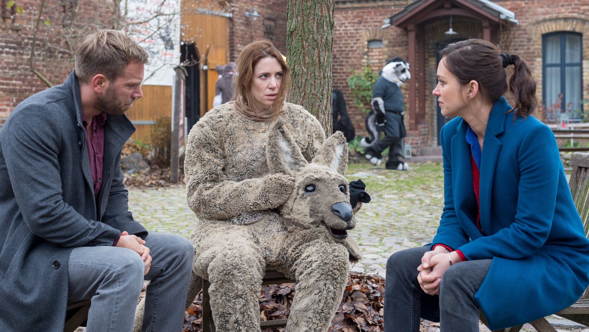 "SOKO Wismar – Der wie ein Wolf tanzt": Lars Pöhlmann (Dominic Boeer) und Karoline Joost (Nike Fuhrmann) sitzen mit Patricia Weiss (Julia Malik) in einem Hof. Sie hat den Kopf ihres Känguru-Kostüms abgesetzt und auf ihren Schoß gelegt und blickt unsicher zu Karoline. Im Hintergrund erkennt man einige Furries (Komparsen).