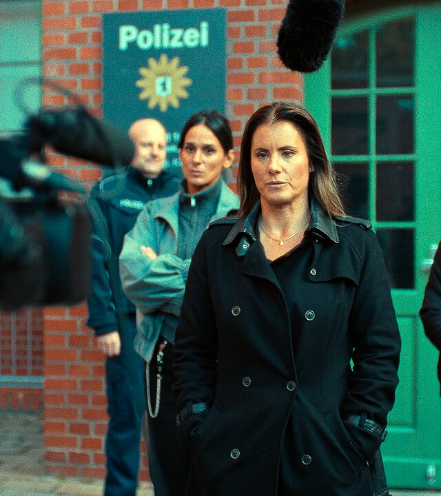 "Jenseits der Spree - Kontrollverlust": Ein Polizist (Komparse), Mavi Neumann (Aybi Era), Katharina Koblinski (Elisabeth Baulitz) und Robert Heffler (Jürgen Vogel) stehen vor dem Eingang eines Polizeigebäudes. An den Seiten im Anschnitt befinden sich eine Kamera und ein Mikrofon. Katharina Koblinski steht vor der Kamera, die anderen stehen hinter ihr. An der Wand neben der Eingangstür hängt das Schild der Dienststelle.