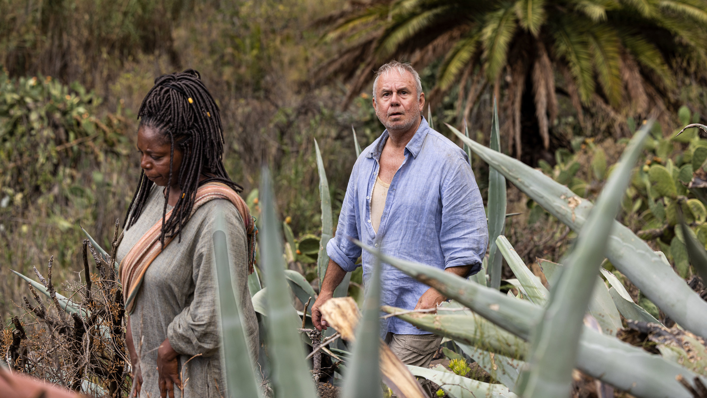 "Endlich Witwer - Über alle Berge": Hinter der Vegetation der Insel folgt Weiser (Joachim Król) erneut Soleil (Dela Dabulamanzi). Während ihr Blick nach unten geht, schaut Weiser beunruhigt und fragend auf.