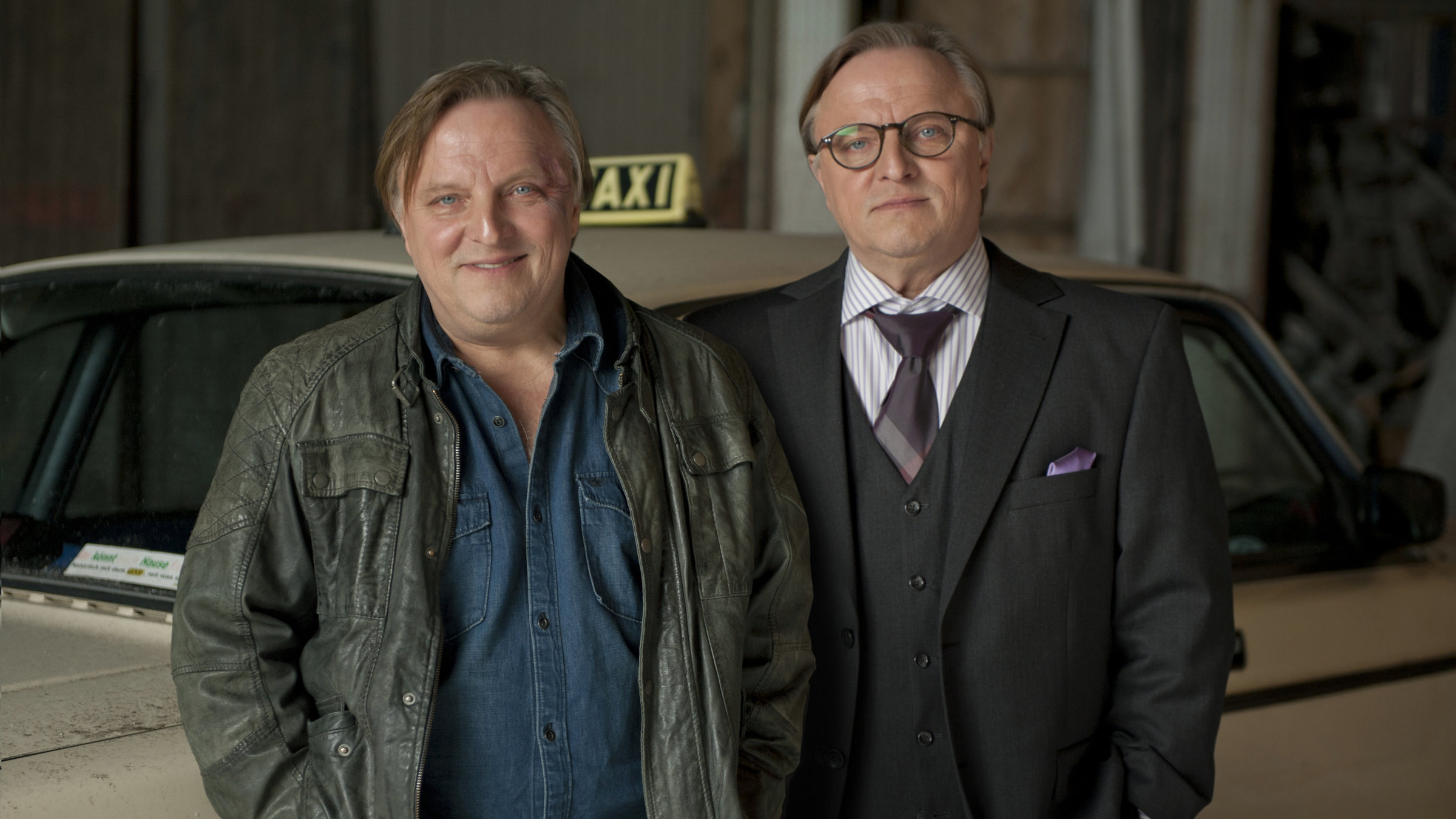 „Die Lichtenbergs – zwei Brüder, drei Frauen und jede Menge Zoff“: Jochen Lichtenberg (Axel Prahl) und Christian Lichtenberg (Axel Prahl), beider sehr unterschiedlich gekleidet, stehen nebeneinander in vor einem Taxi.