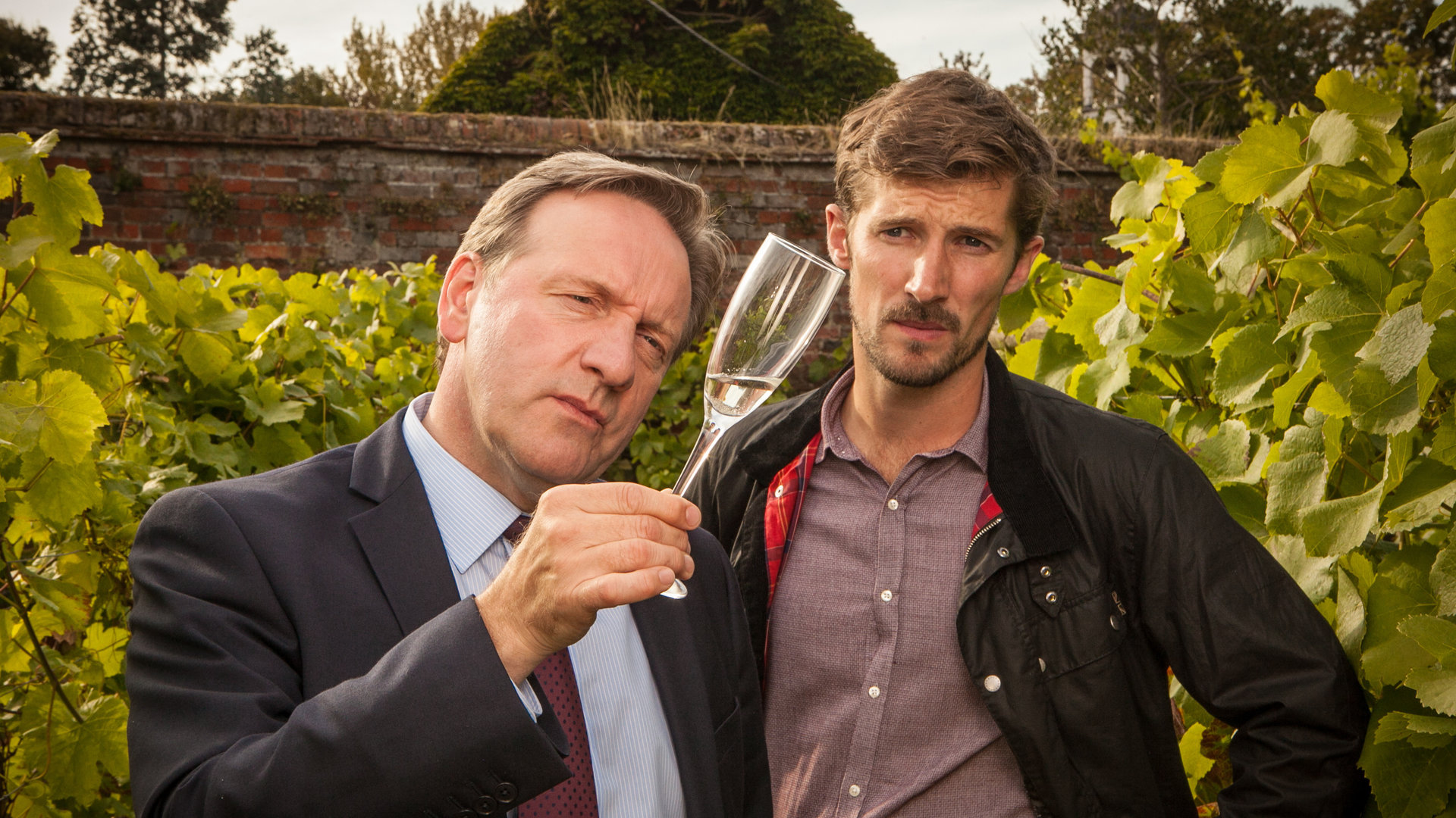 "Inspector Barnaby - Ein letzter Tropfen": DCI Barnaby (Neil Dudgeon) und Charlie Nelson (Gwilym Lee) betrachten skeptisch ein Sektglas, das Barnaby in der Hand hält.