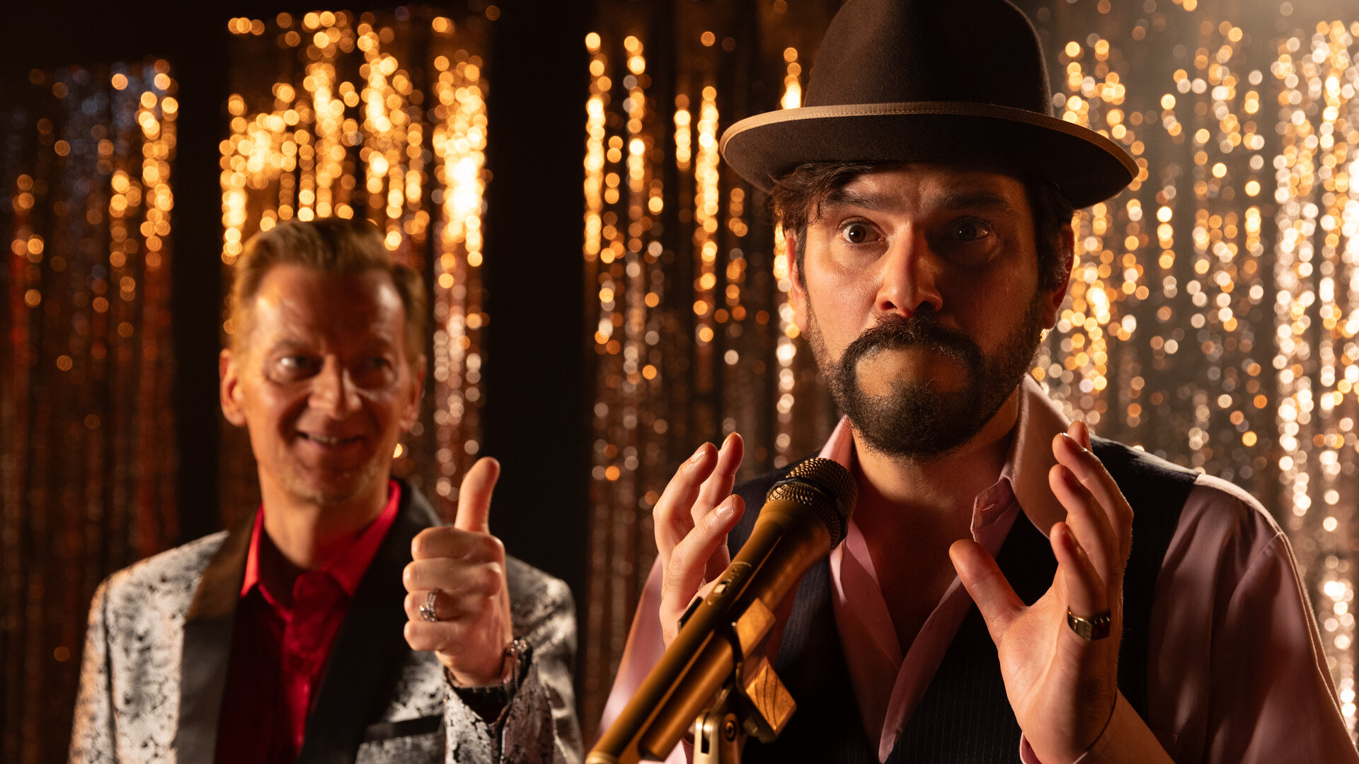 "Rembetis - Die Geisterjäger - Don't Worry Be Happy": Florian Goldesel (Michael Kessler) und Paris (Jasin Challah) stehen gemeinsam auf einer Bühne. Während Florian Goldesel einen Daumen hoch zeigt, steht Paris mit schockiertem Blick und verschlossenem Mund vor einem Mikrofon. Im Hintergrund hängt ein golden glitzernder Lamettavorhang.