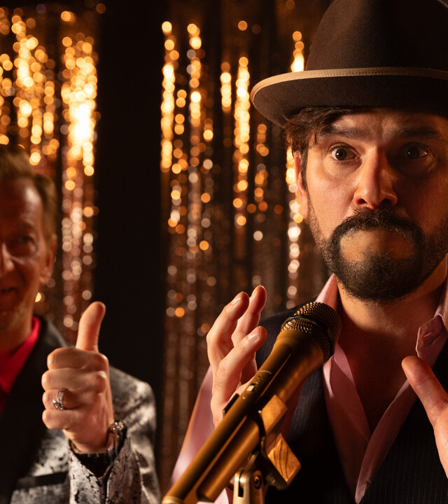"Rembetis - Die Geisterjäger - Don't Worry Be Happy": Florian Goldesel (Michael Kessler) und Paris (Jasin Challah) stehen gemeinsam auf einer Bühne. Während Florian Goldesel einen Daumen hoch zeigt, steht Paris mit schockiertem Blick und verschlossenem Mund vor einem Mikrofon. Im Hintergrund hängt ein golden glitzernder Lamettavorhang.