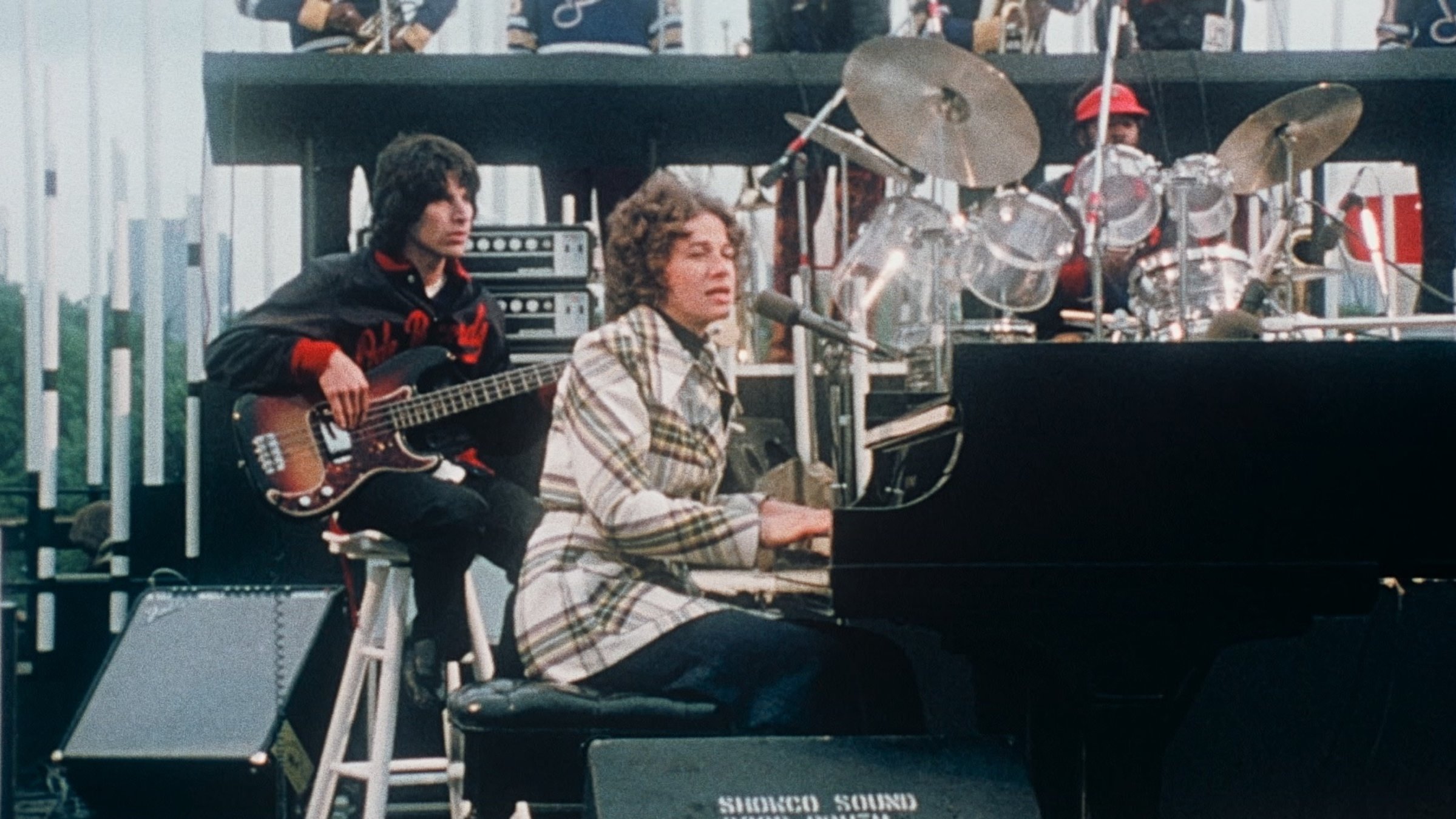 "Carole King: Home Again - Live in Central Park": Die Sängerin Carole King sitzt an einem Flügel auf einer Bühne und singt. Hinter ihr sitzt ein Musiker auf einem Hocker und spielt E-Gitarre. Dahinter ein weiterer Musiker an einem gläsernen Schlagzeug.