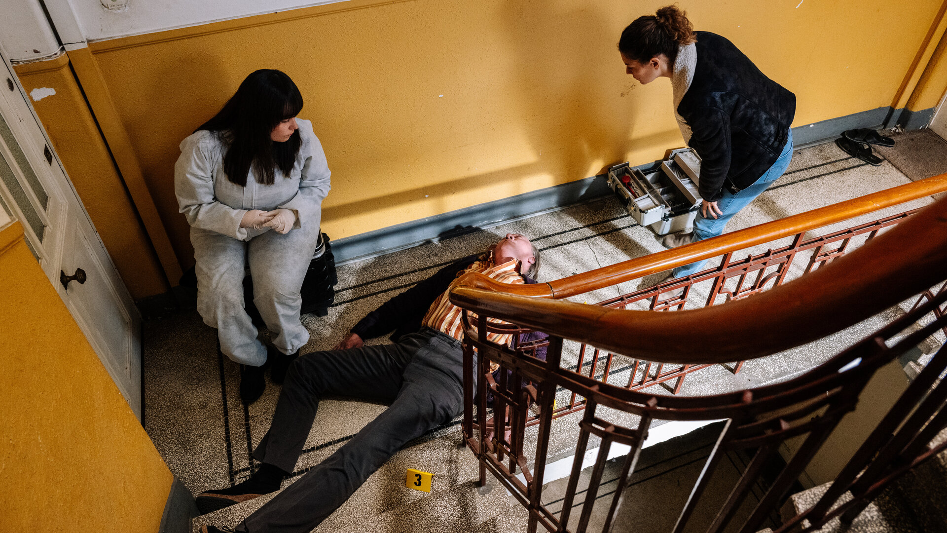 "SOKO Köln - Adlerauge": Blick von schräg oben: Dr. Westphal (Yulia Yáñez Schmidt.) und Lilly Funke (Tatjana Kästel) befinden sich in einem gelb-weiß gestrichenen Treppenhaus und blicken auf den toten Peter Canapi (Stephan Böttcher), der auf dem Rücken auf dem Boden liegt. Dr. Westphal trägt einen weißen Overall und sitzt vor einer weißen Wohnungstür. Lilly Funke steht auf der anderen Seite von Peter Canapi und beugt sich leicht über ihn.
