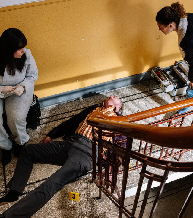 "SOKO Köln - Adlerauge": Blick von schräg oben: Dr. Westphal (Yulia Yáñez Schmidt.) und Lilly Funke (Tatjana Kästel) befinden sich in einem gelb-weiß gestrichenen Treppenhaus und blicken auf den toten Peter Canapi (Stephan Böttcher), der auf dem Rücken auf dem Boden liegt. Dr. Westphal trägt einen weißen Overall und sitzt vor einer weißen Wohnungstür. Lilly Funke steht auf der anderen Seite von Peter Canapi und beugt sich leicht über ihn.