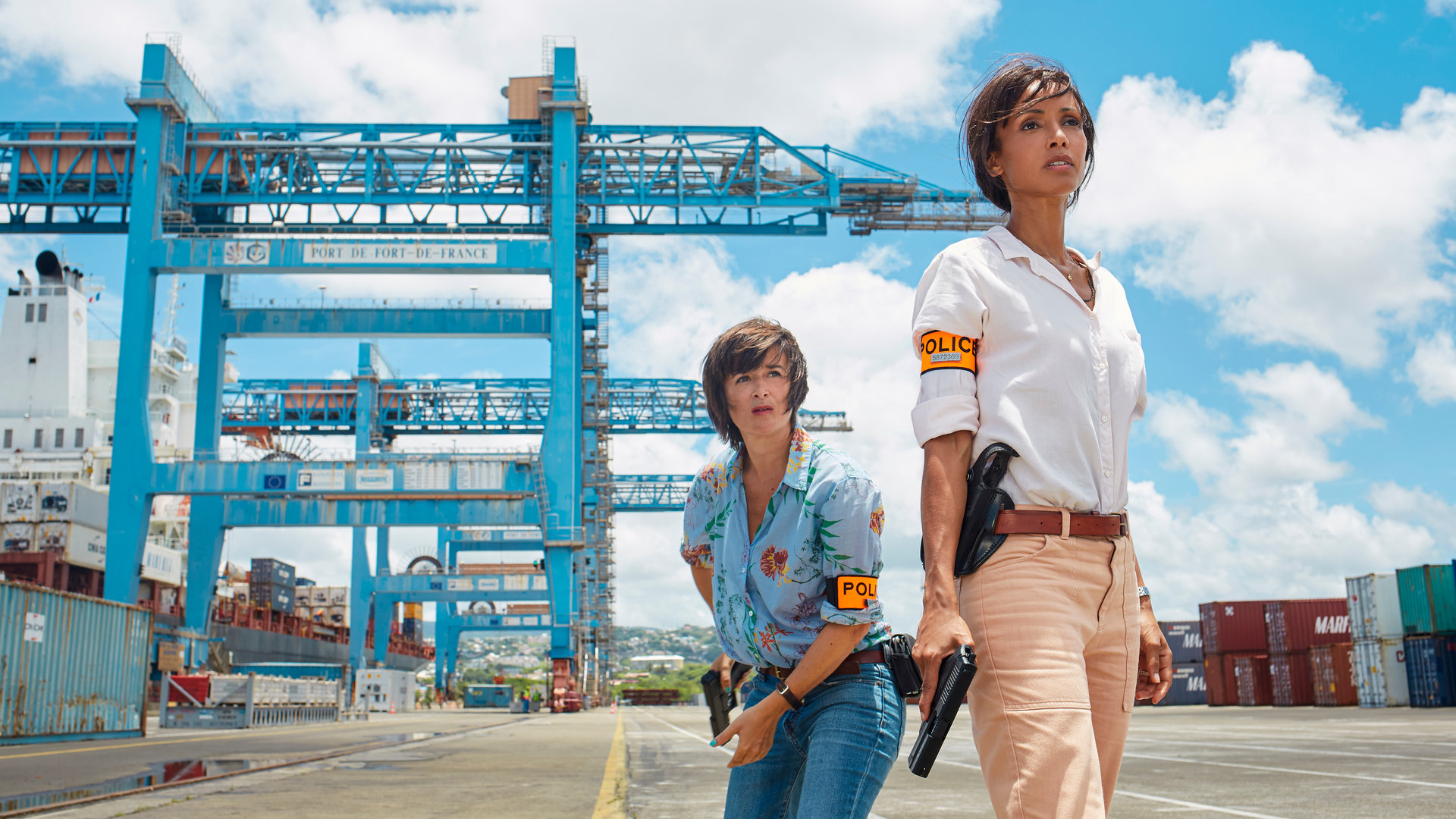 "Deadly Tropics - Die Frau seines Lebens": In einem Container-Hafen. Gaëlle Crivelli (Béatrice de la Boulaye) steht hinter Mélissa Sainte-Rose (Sonia Rolland). Beide schauen ernst in die Ferne und halten dabei Waffen in der Hand.