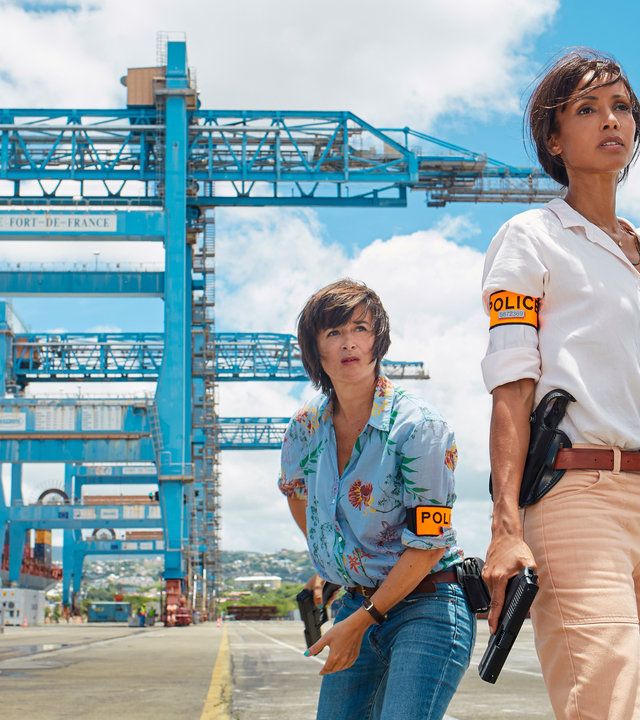 "Deadly Tropics - Die Frau seines Lebens": In einem Container-Hafen. Gaëlle Crivelli (Béatrice de la Boulaye) steht hinter Mélissa Sainte-Rose (Sonia Rolland). Beide schauen ernst in die Ferne und halten dabei Waffen in der Hand.