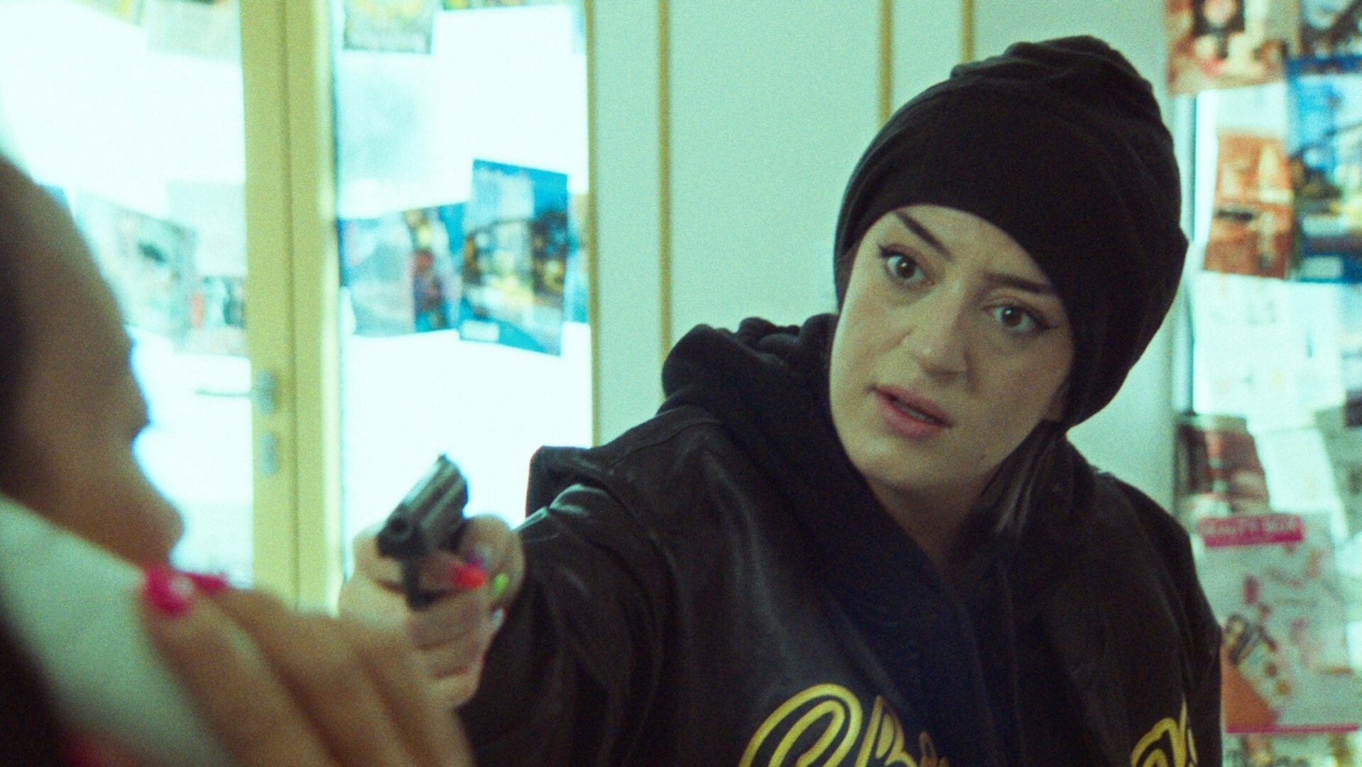 "Doppelhaushälfte – Überfall": Im Kosmetikstudio zielt Räuberin Becky (Parshad Esmaeli) mit einer Pistole seitlich nach vorne. Ihr Gesicht ist angespannt und sie trägt schwarze Kleidung.