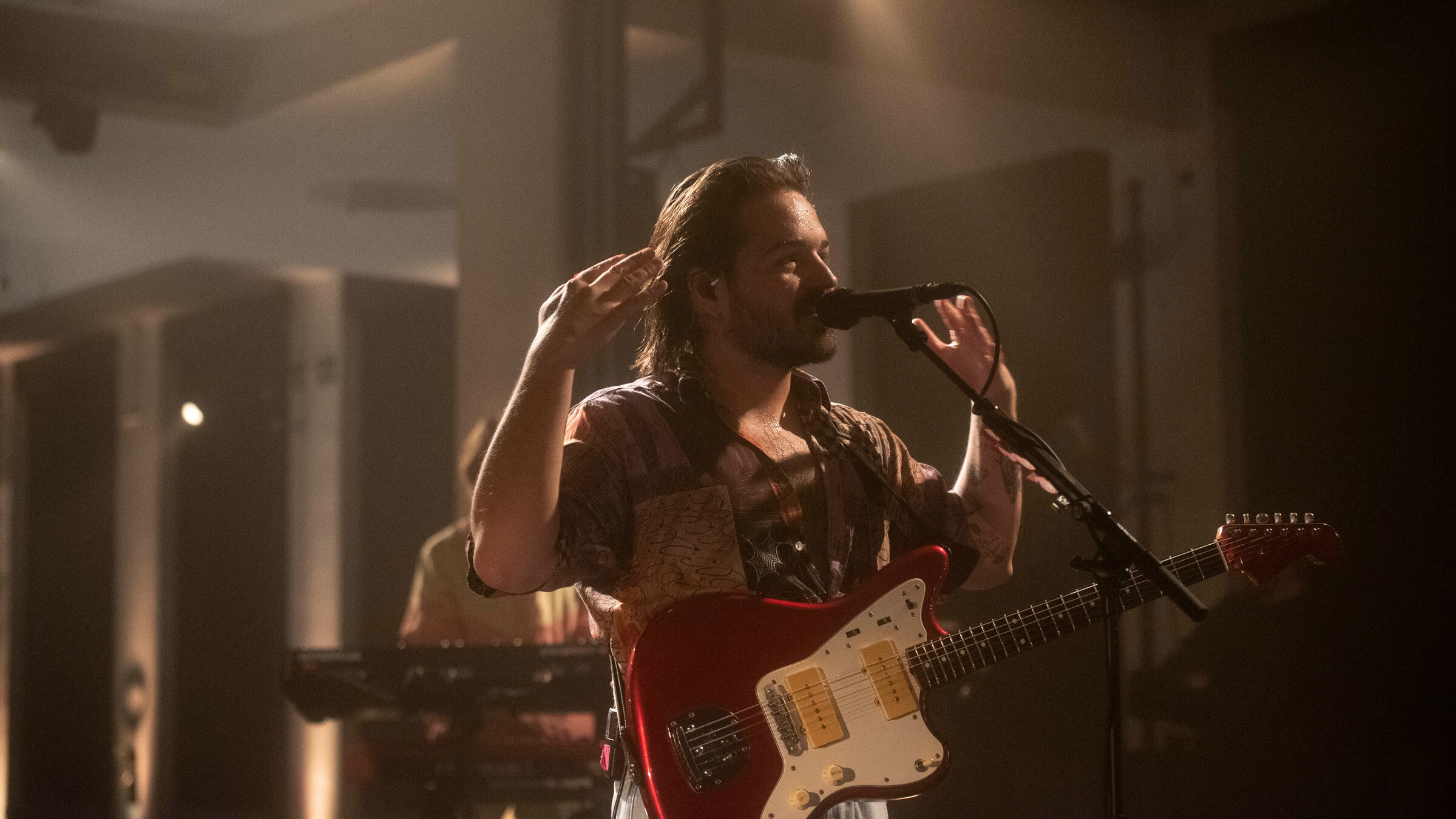 "Pop Around@Bauhaus: Milky Chance": Der Musiker Clemens Rehbein steht auf einer Bühne vor einem Mikrofonständer und singt in das Mikrofon. Die Arme hat er erhoben. Er trägt eine E-Gitarre um die Schulter. Im Hintergrund - fast verdeckt durch die vordere Person - erkennt man eine Person an einem Keyboard.