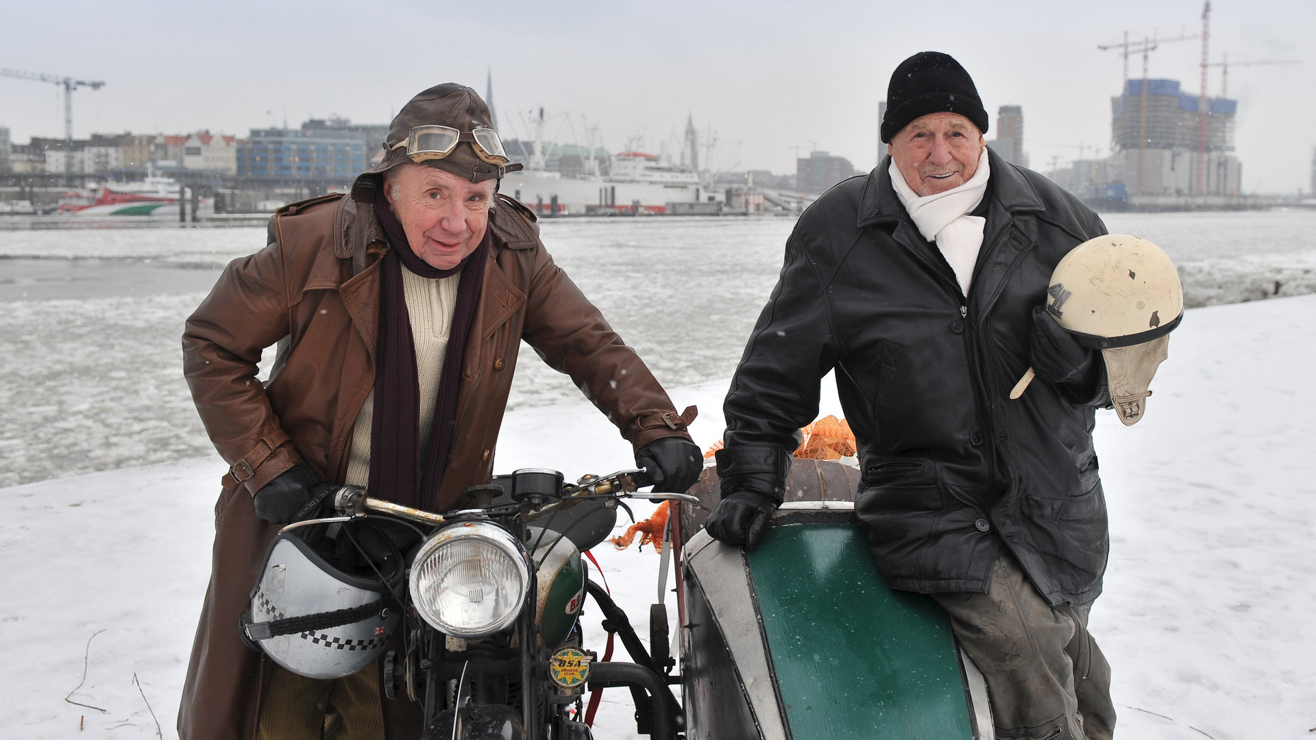 "Notruf Hafenkante - Alte Freunde": Arno (Herbert Köfer) und Kuddel (Horst Sachtleben) stehen links und rechts neben einem Motorrad mit Beiwagen am verschneiten Hafen und lächeln in die Kamera.